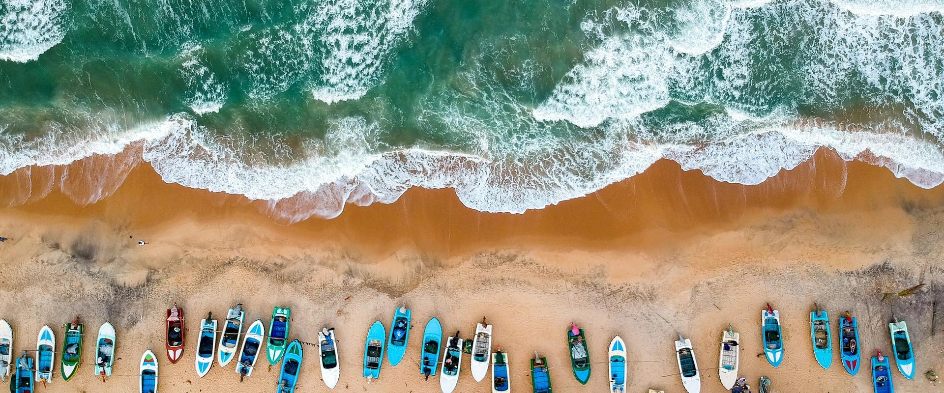 Aerial view of colorful fishing boats lined up on golden sand at Arugam Bay, Sri Lanka, with turquoise Indian Ocean waves crashing onto the beach.