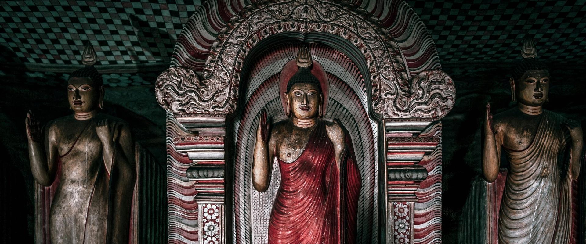 Three ancient Buddha statues inside Dambulla Cave Temple, Sri Lanka, with ornate red-and-white arched shrine and painted ceiling