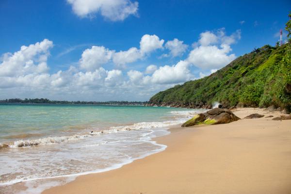 Unawatuna Beach in Sri Lanka with golden sand, turquoise Indian Ocean waves, rocky shoreline, and lush green hillside under a bright blue sky with scattered clouds.