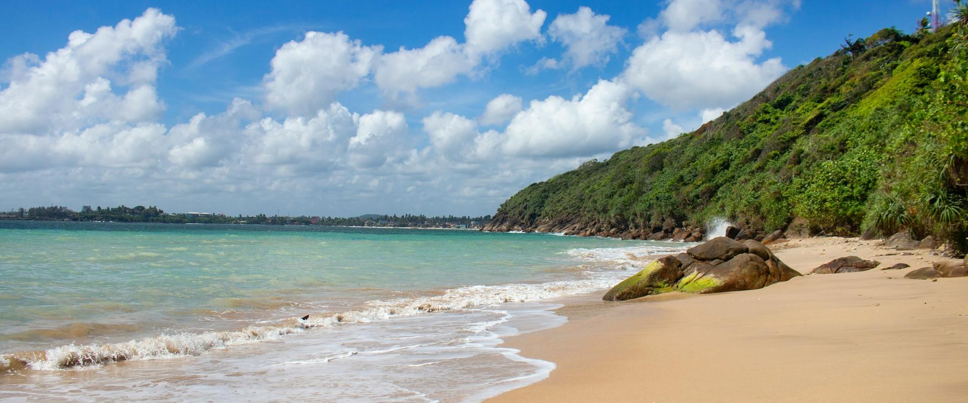 Unawatuna Beach in Sri Lanka with golden sand, turquoise Indian Ocean waves, rocky shoreline, and lush green hillside under a bright blue sky with scattered clouds.