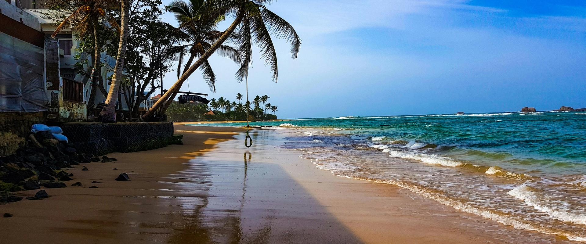 A rope swing hanging from a leaning palm tree on a golden sandy beach with turquoise waves, guesthouses lining the shore and rocky outcrops visible in the distance, Hikkaduwa, Sri Lanka