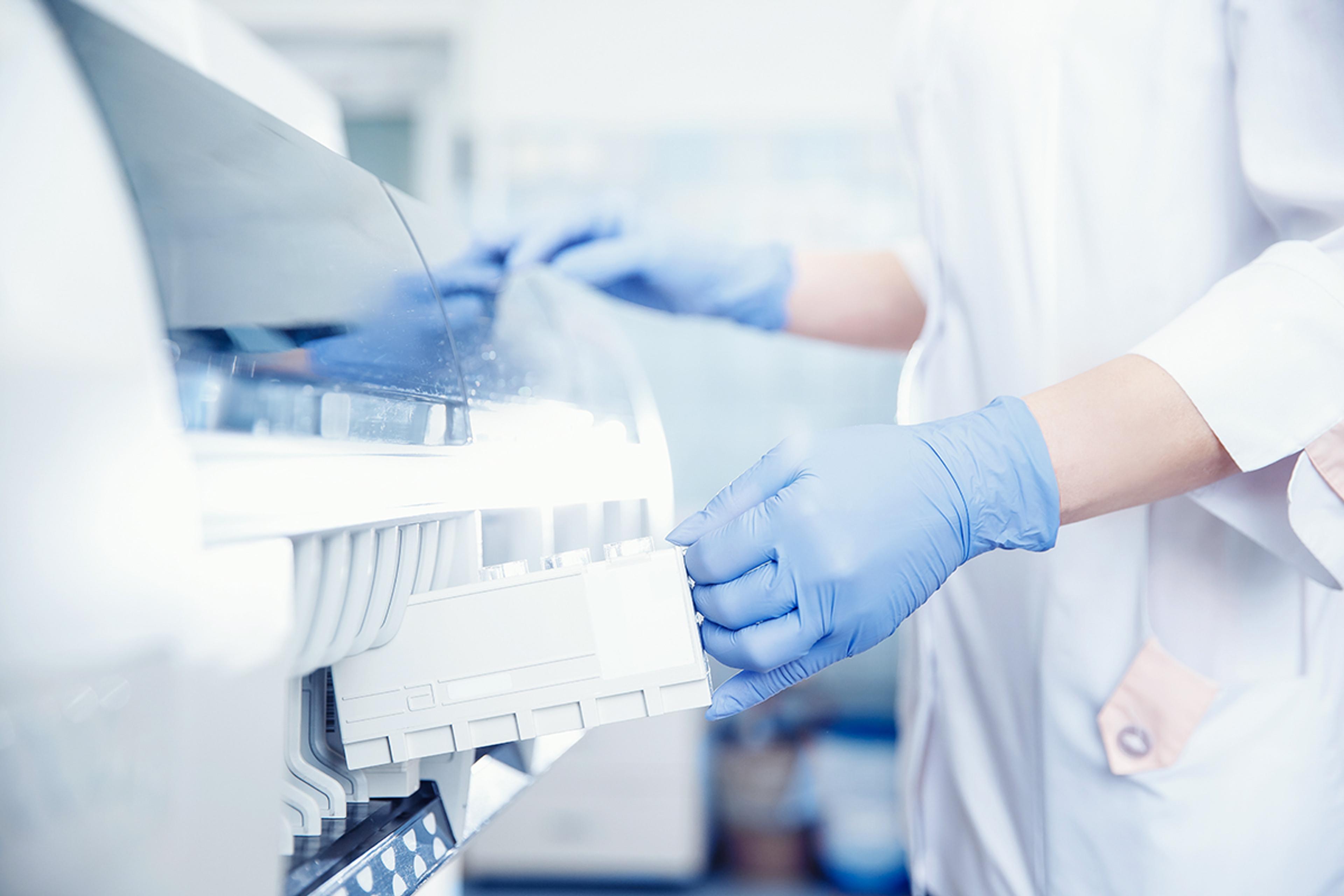 Gloved hands loading sample tubes into an automated laboratory analyser, with a clinician in a lab coat in the background.
