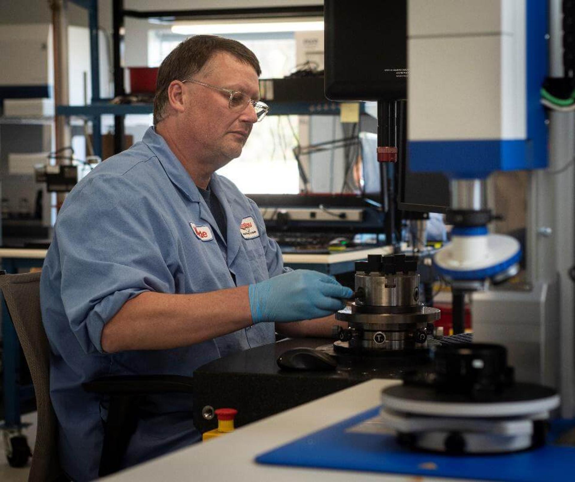 Engineer at work at G&H | Stingray - A male technician wearing a blue lab coat, safety glasses, and blue gloves is working with precision components at a workstation in a manufacturing or engineering facility. He is focused on inspecting or assembling a metallic cylindrical part using a high-precision measurement or alignment device. The environment is clean and equipped with advanced technical equipment.