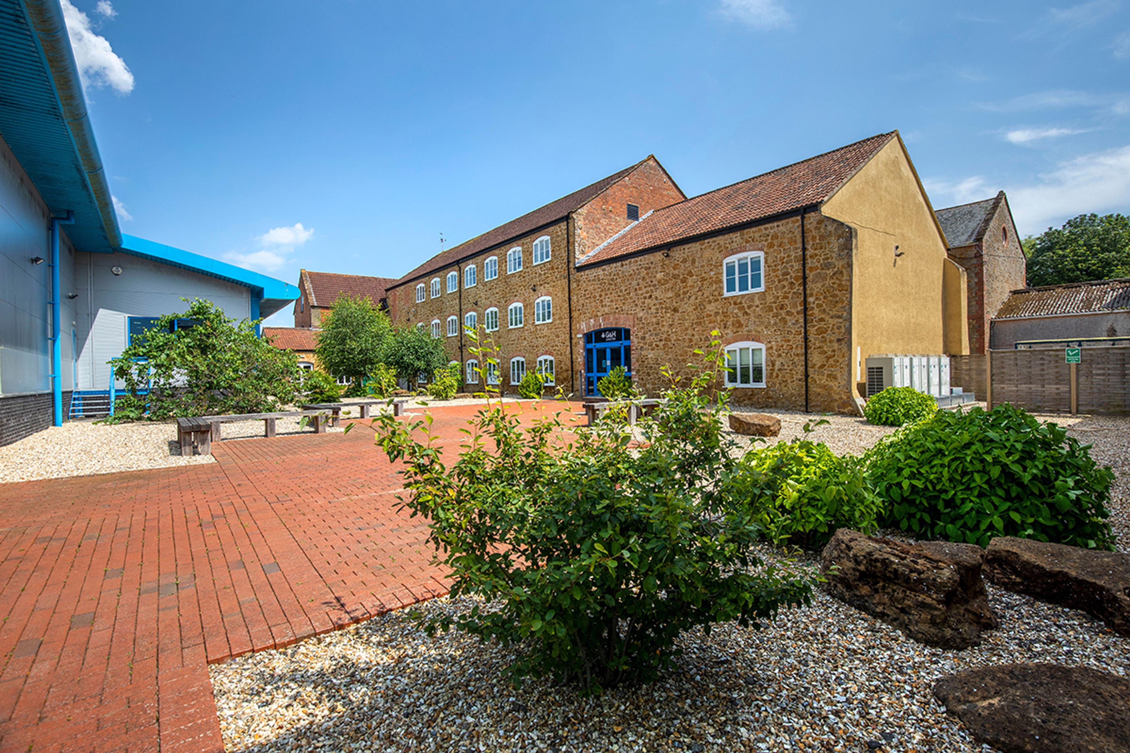 Courtyard view of G&H's Dowlish Ford facility showing a mix of historic stone buildings and modern industrial units under a clear blue sky, with landscaped red brick pathways, benches, and lush green shrubbery.