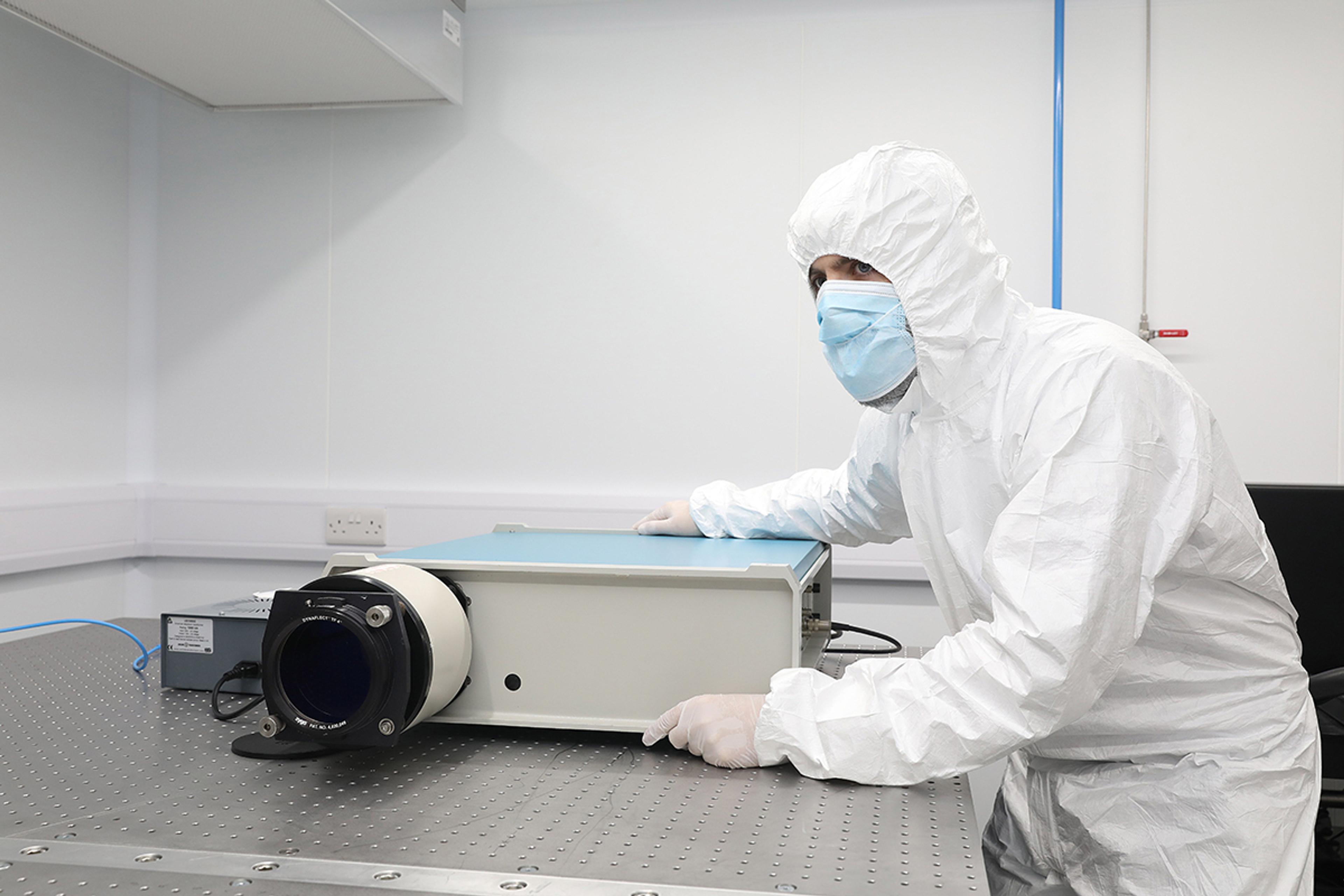 Engineer wearing a full-body cleanroom suit, gloves, and face mask operating a large scientific optical instrument on a vibration-damped optical table. The lab setting is sterile and brightly lit, designed for precision optical or photonics work.