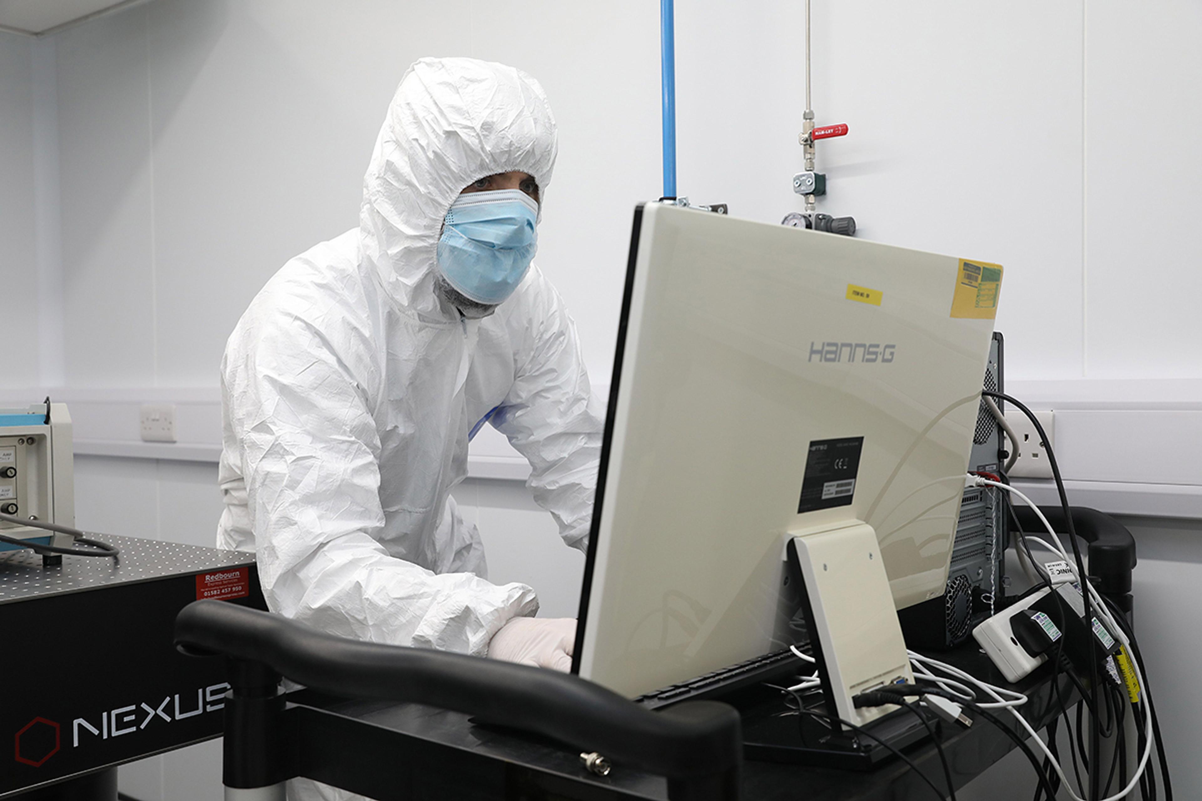 Technician in full cleanroom attire, including a white hooded suit, face mask, and gloves, working at a computer station inside a controlled laboratory environment. The workspace features scientific equipment and a Hanns.G monitor, with visible cable connections and a NEXUS optical table.