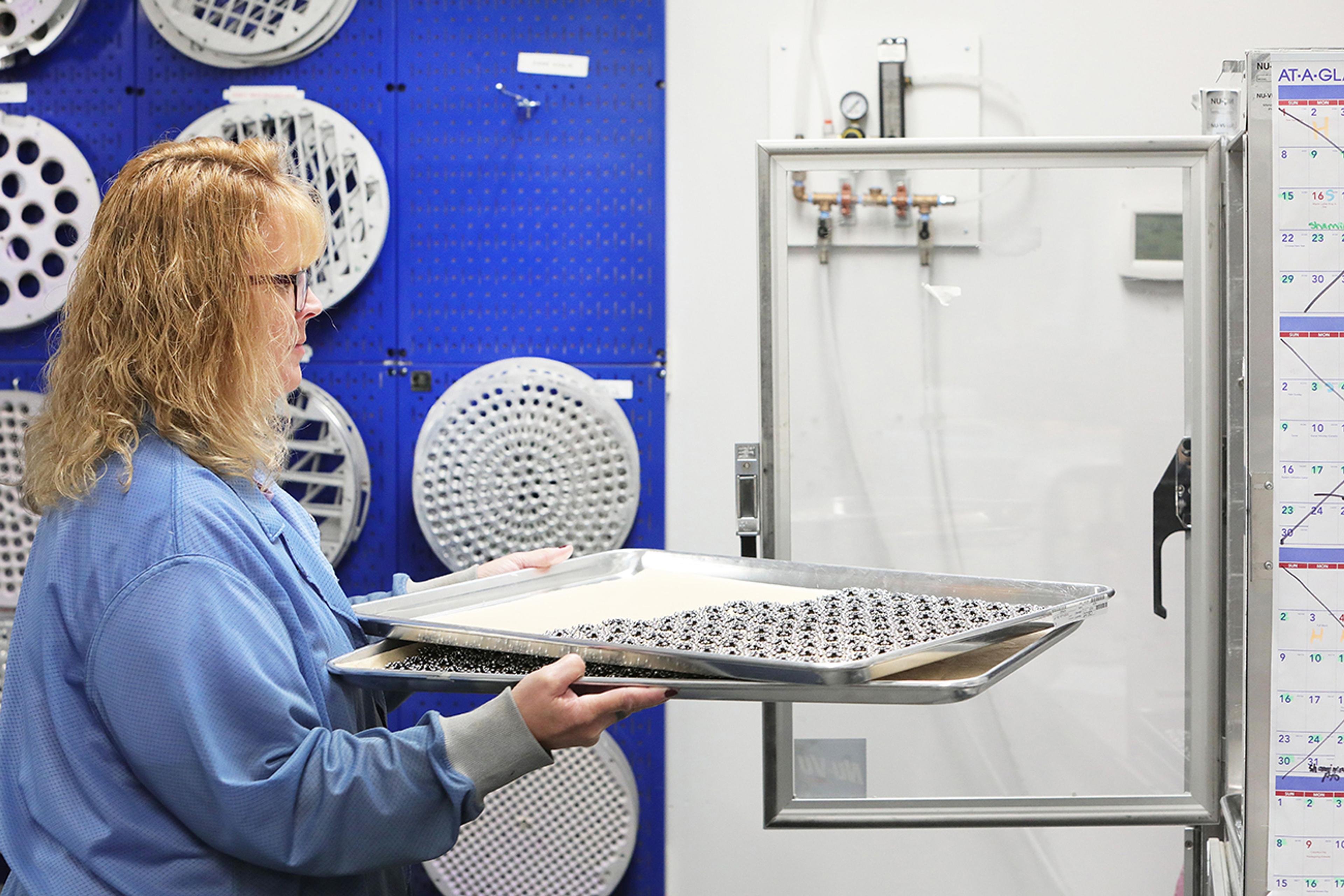 Polymer optics, A woman in a blue lab coat is placing a metal tray filled with small black optical components into a transparent drying or cleanroom chamber. She is working in a clean manufacturing environment, with a blue tool board holding various metal templates or fixtures in the background. A wall calendar is partially visible on the right side of the image, adding context to the workspace