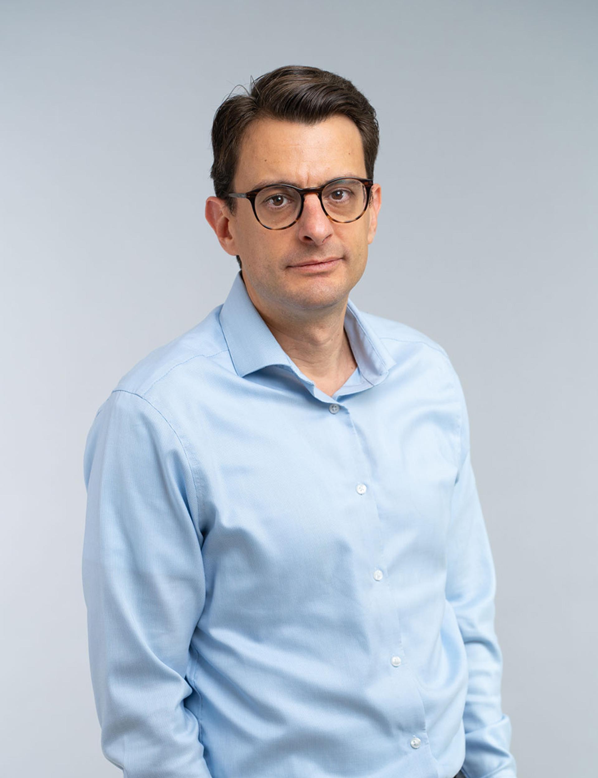 Stratos Kehayas, PhD, G&H President of Photonics - A professionally dressed man in a  light blue shirt stands against a light grey background. He has short dark hair, wears glasses, and has a neutral expression with his hands in his pockets.