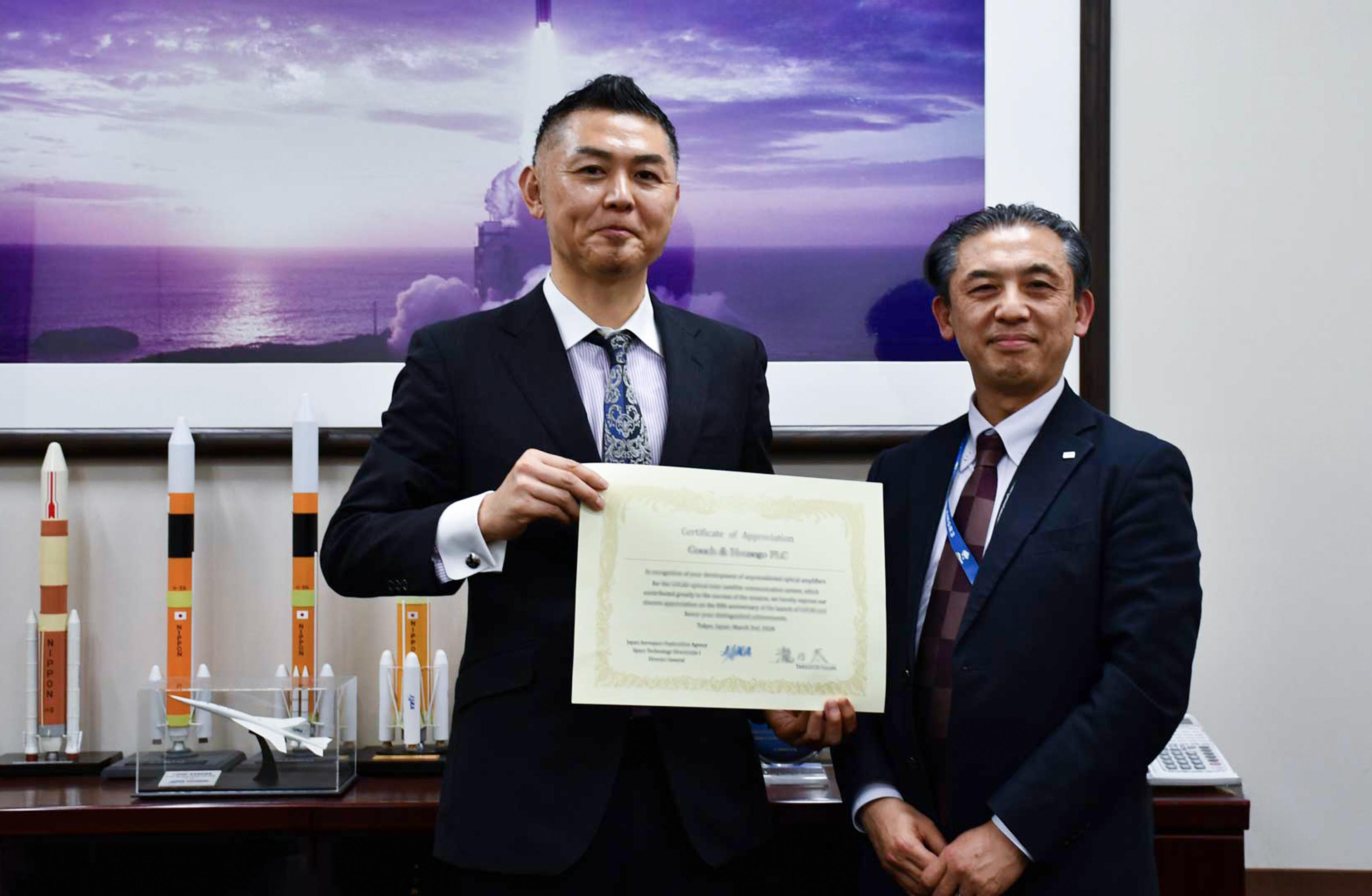 Two suited G&H representatives display a JAXA Letter of Appreciation in recognition of the company’s contribution to the LUCAS Optical Data Relay Satellite Communication System, with space launch imagery and rocket models in the background.