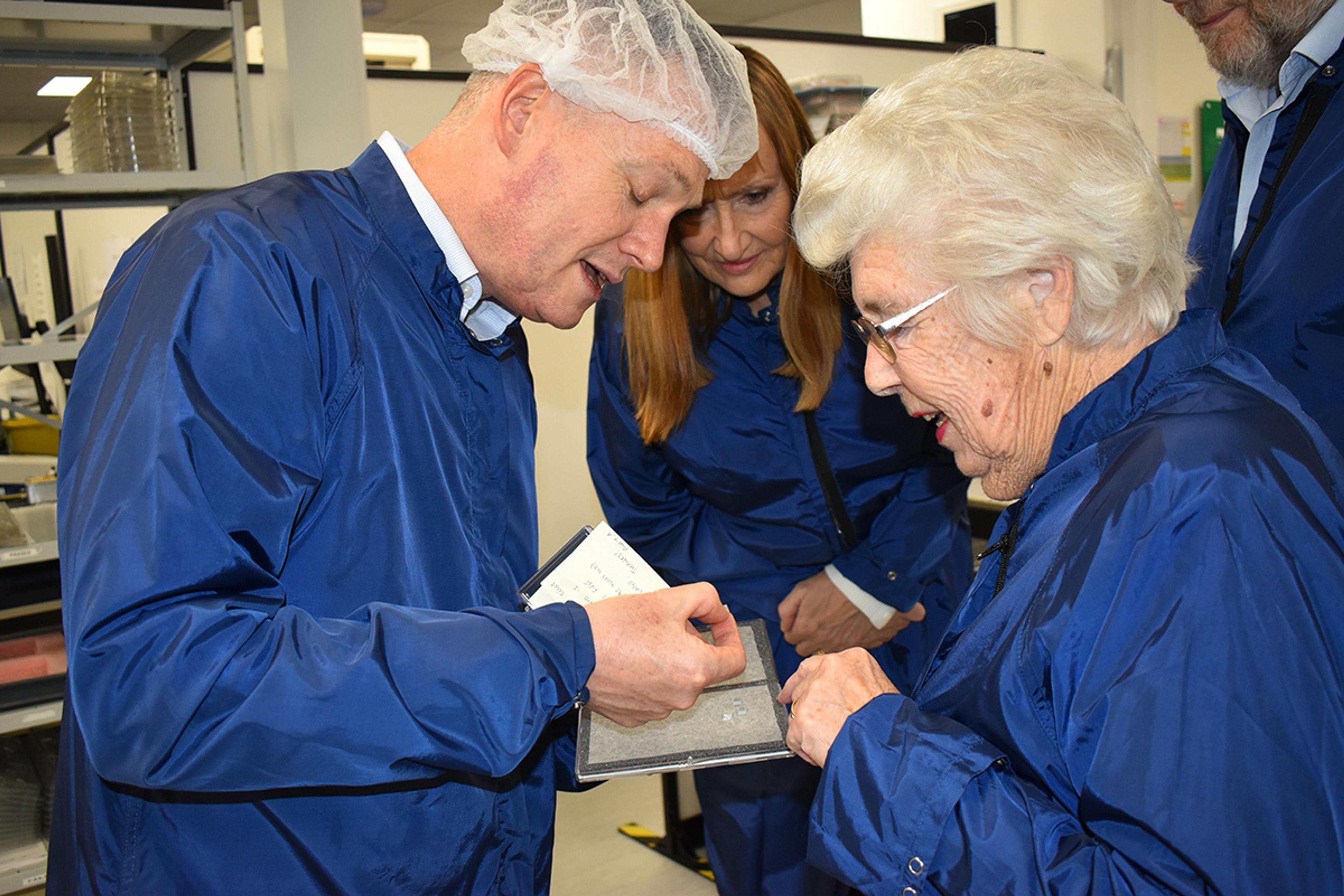 Toby Reid (Left), Product Management Director, G&H Torquay & Norma Knapp (Right) In a cleanroom environment, a man wearing a hairnet and blue lab coat shows a small component or sample to an elderly woman also in a blue lab coat. He holds the item carefully on a tray or notebook while explaining its features. Another woman and man in similar attire observe closely from behind, all appearing engaged and interested. The setting suggests a guided tour or educational visit within a high-tech or manufacturing facility.
