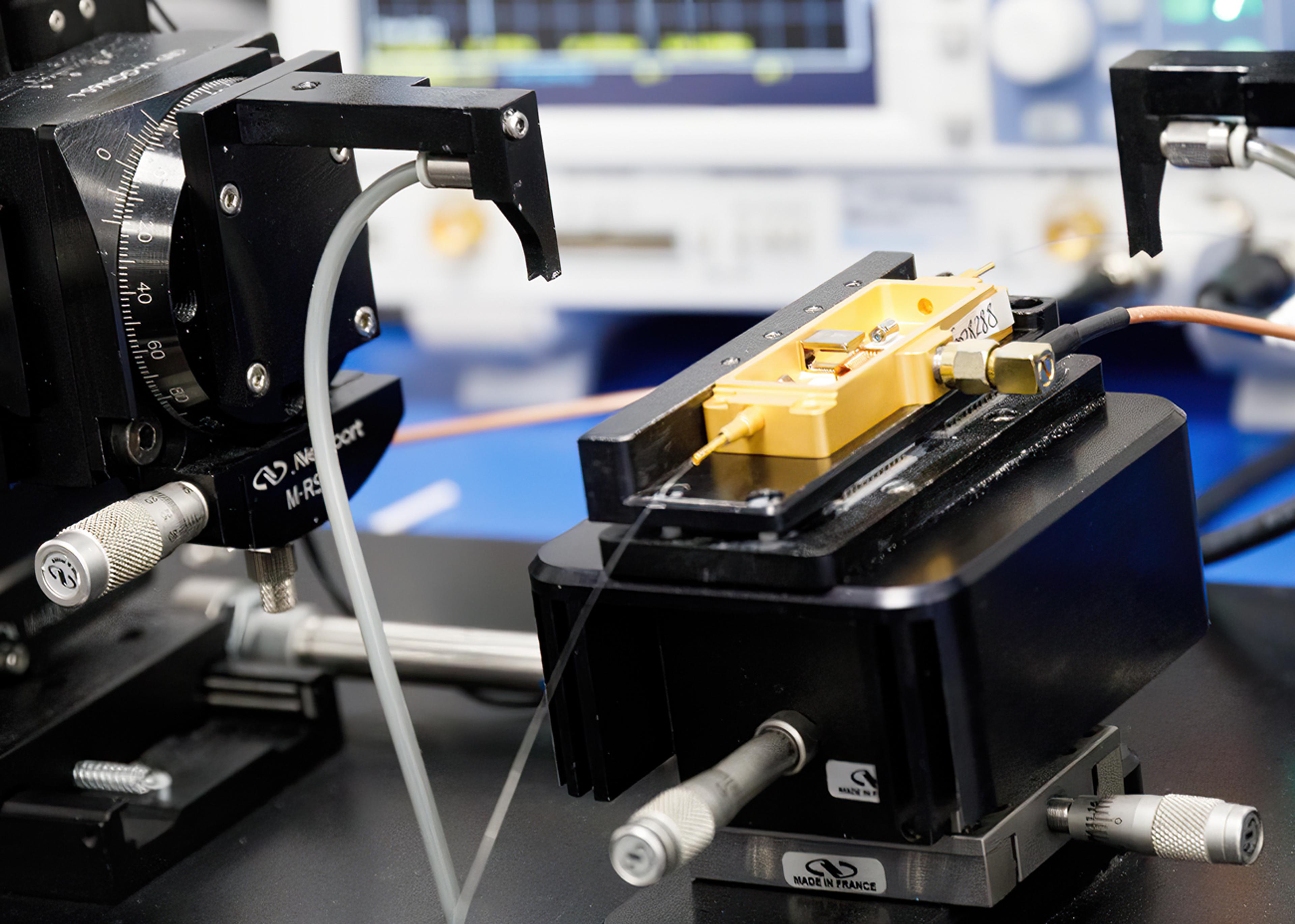 Gold photonics module mounted on a precision positioning stage within an optical test setup, with cables connected and measurement equipment blurred in the background.