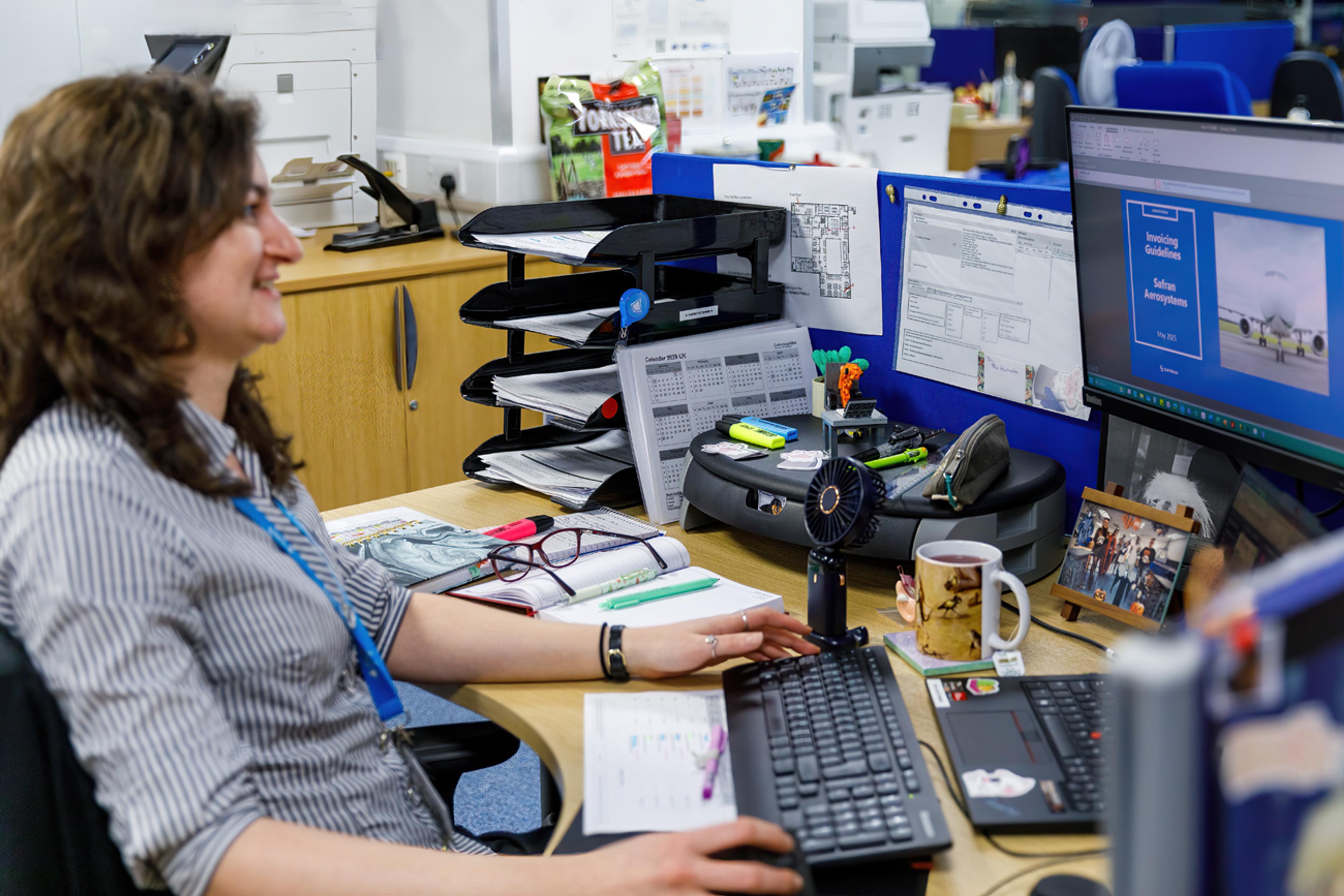 Office employee wearing a lanyard works at a desk with dual monitors, paperwork trays and stationery, with an “Invoicing Guidelines” document open on the screen.