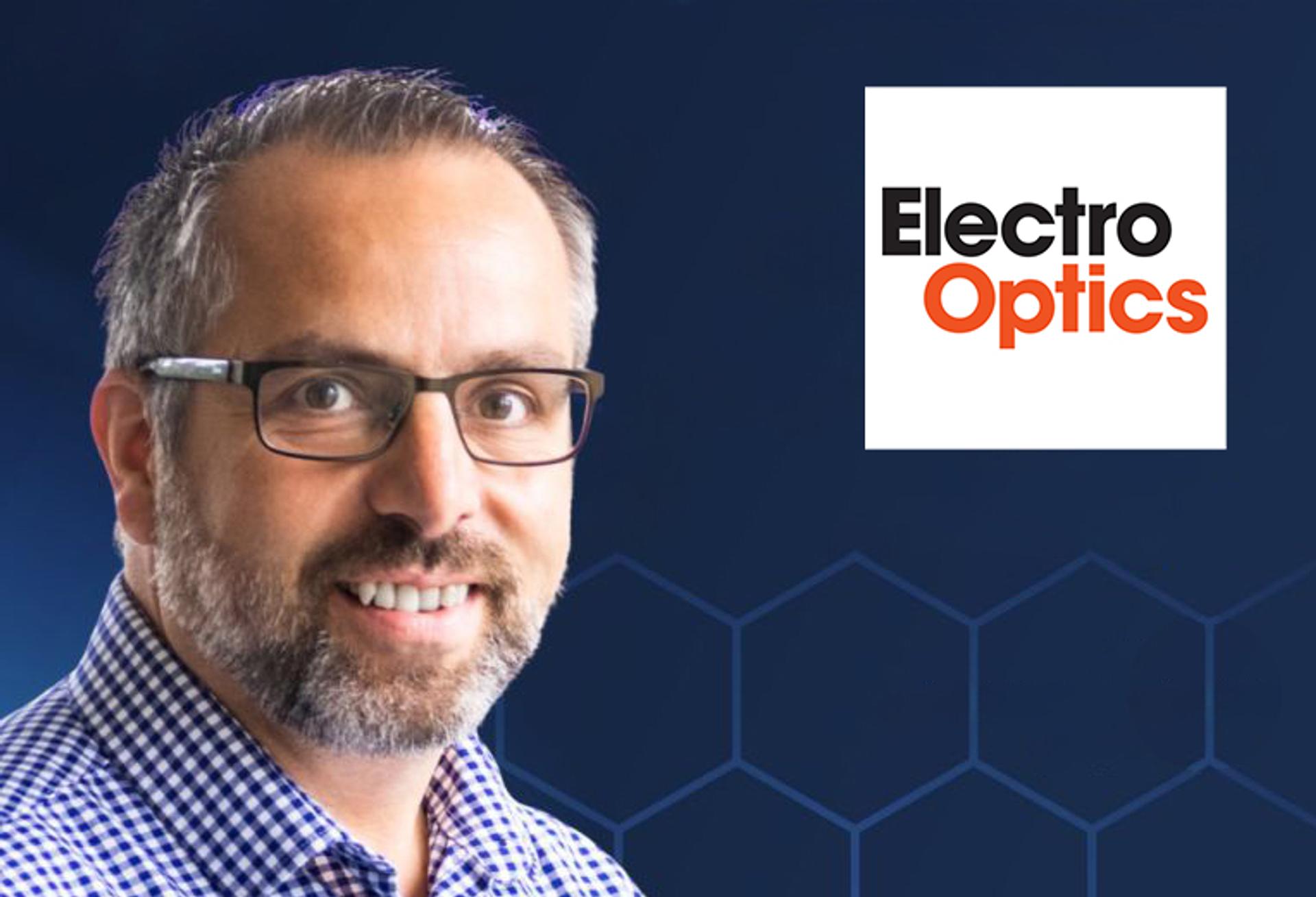 Matt Hall. Portrait of a man wearing glasses and a blue checkered shirt, smiling against a dark blue background with a hexagonal pattern. The Electro Optics logo appears in the top right corner.