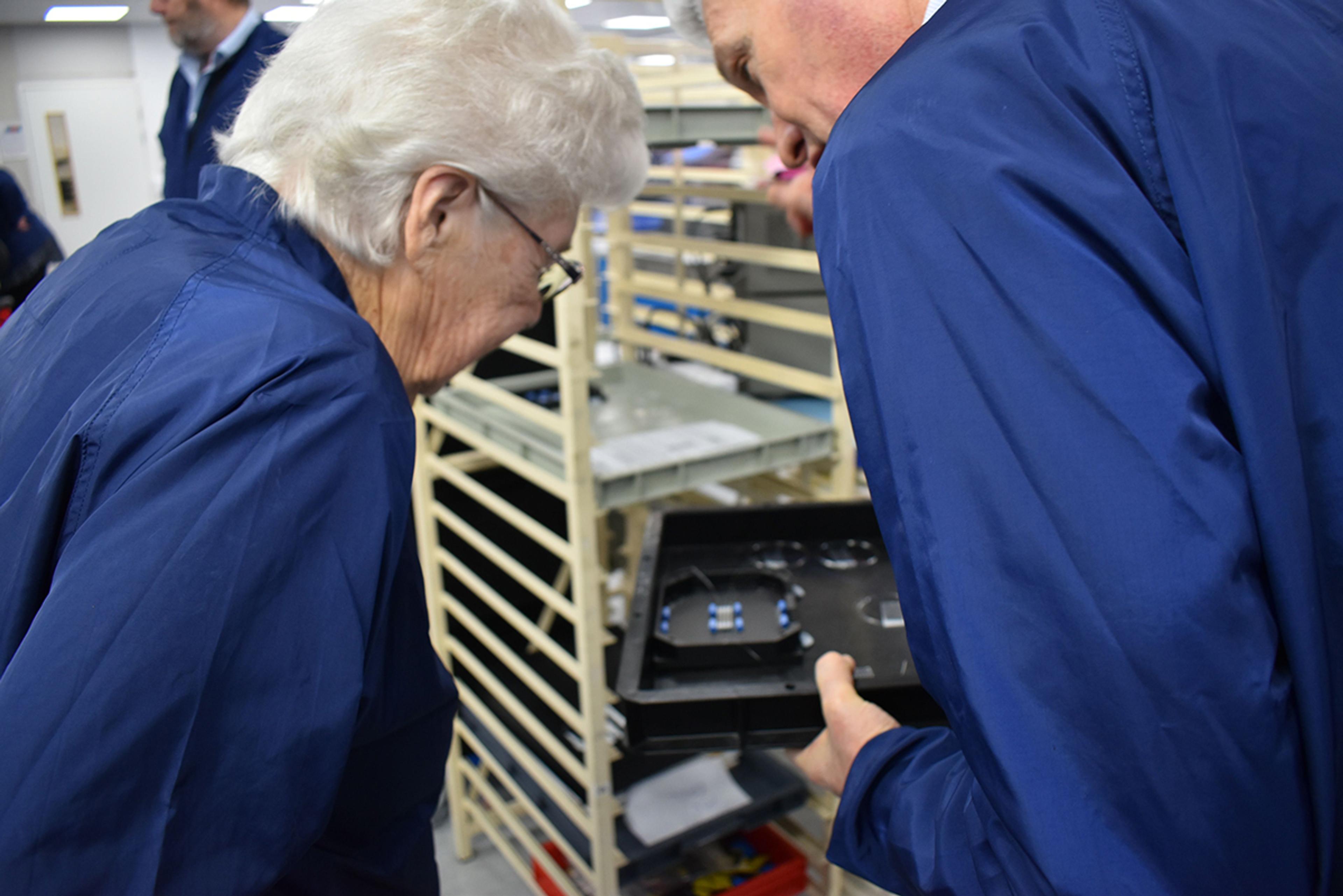 Norma Knapp (L) & Toby Reid (R), Product Management Director, G&H Torquay. Two people wearing blue lab coats examine a black tray containing optical or electronic components inside a manufacturing or lab facility. The elderly woman on the left, with white hair and glasses, leans in closely to view the items, while the man on the right holds the tray and appears to be explaining or showcasing its contents. In the background, shelves and other individuals in similar attire suggest an organized technical environment, possibly a tour of a production or research facility.