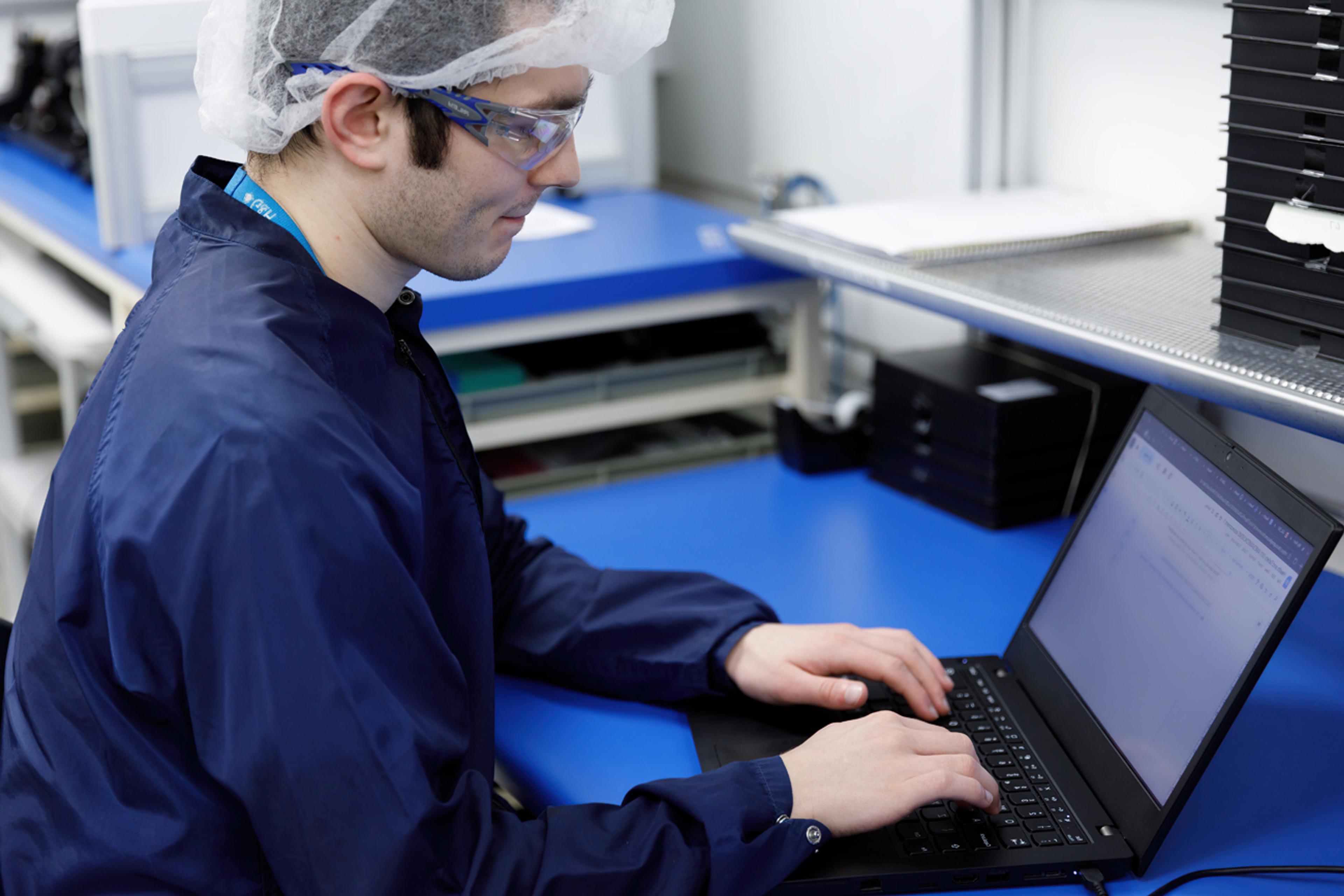 G&H technician wearing cleanroom attire, safety glasses, and a hair cover, working at a blue laboratory bench while typing on a laptop in a controlled manufacturing environment.