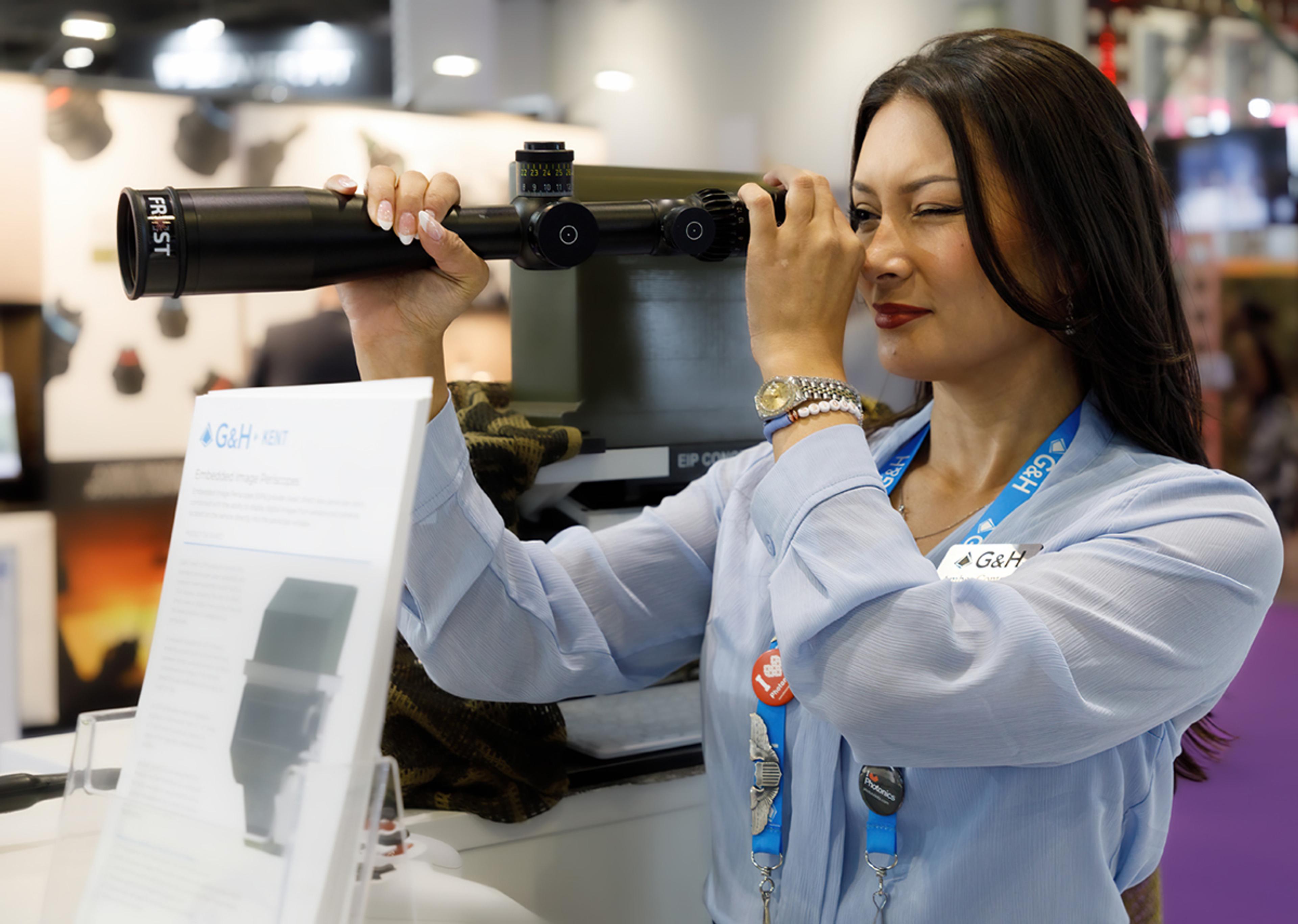 G&H team member at an exhibition looks through a long optical sight or scope on a display stand, wearing a G&H lanyard and name badge.