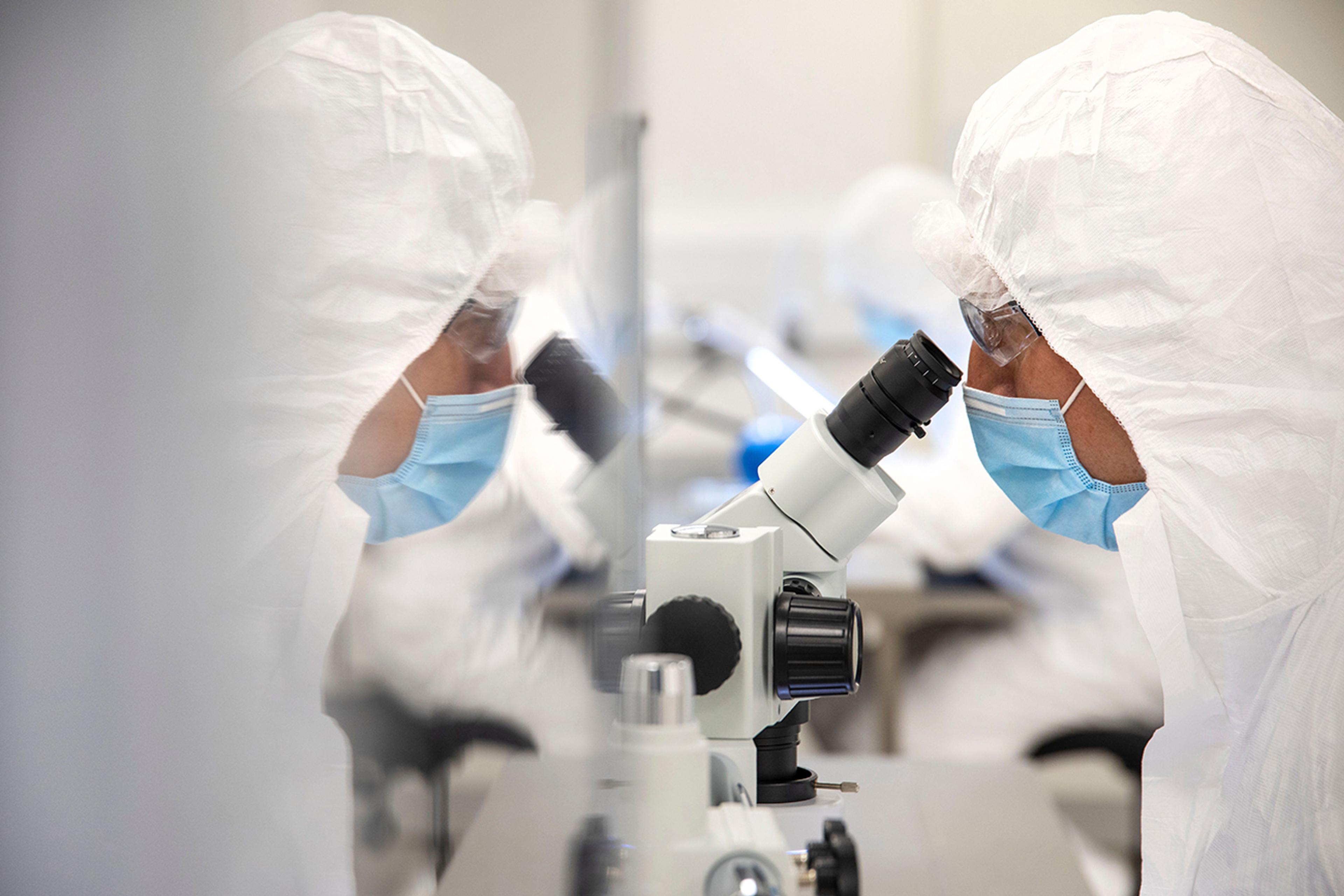 A person wearing a full cleanroom suit, including a face mask and goggles, is looking into a microscope in a sterile laboratory environment. The image shows a clear reflection of the individual and equipment on a glass partition.
