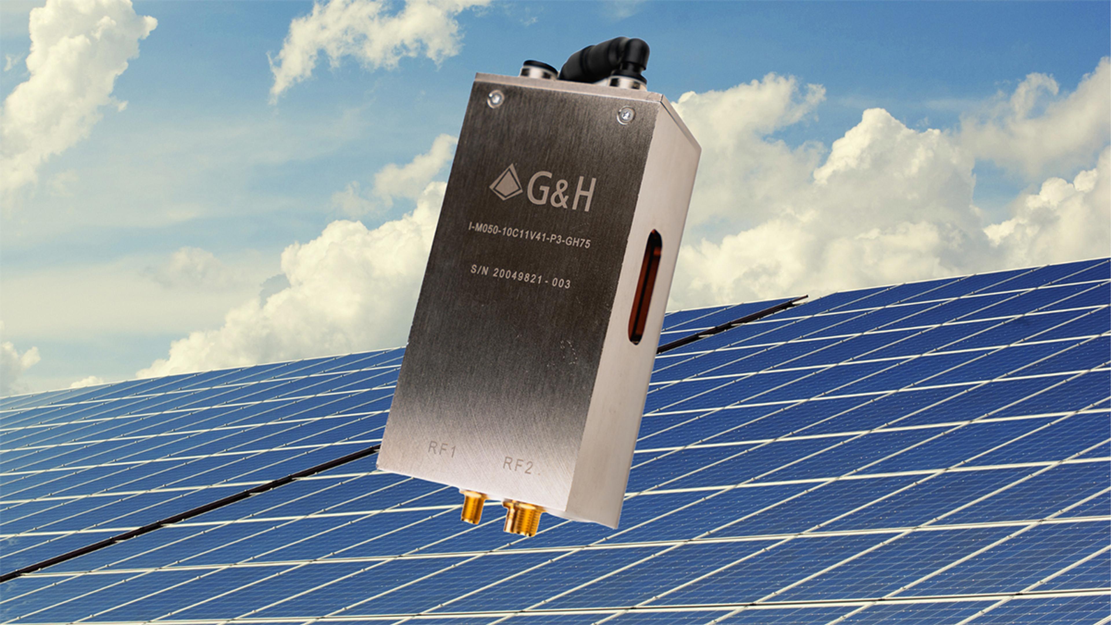 A G&H-branded fiber optic component with engraved serial number is shown prominently in the foreground, angled upward against a bright blue sky with scattered clouds. Behind it lies a large solar panel array stretching across the bottom half of the image, symbolizing clean energy and sustainability. The image represents advanced photonic technology contributing to renewable energy solutions