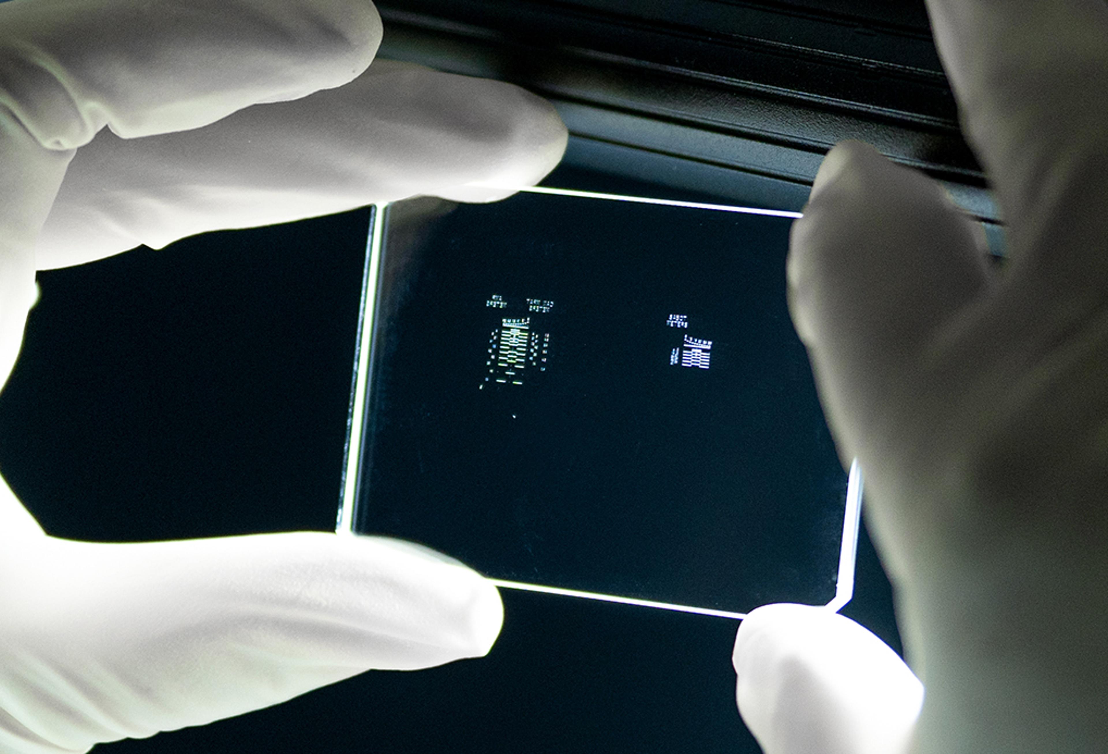 G&H Patterned Dichroic Reticle - A person wearing white gloves holds a transparent optical reticle up to a light source. The rectangular glass plate features fine, high-precision etched patterns and markings visible at its center, used for calibration or alignment in photonics and optical systems.