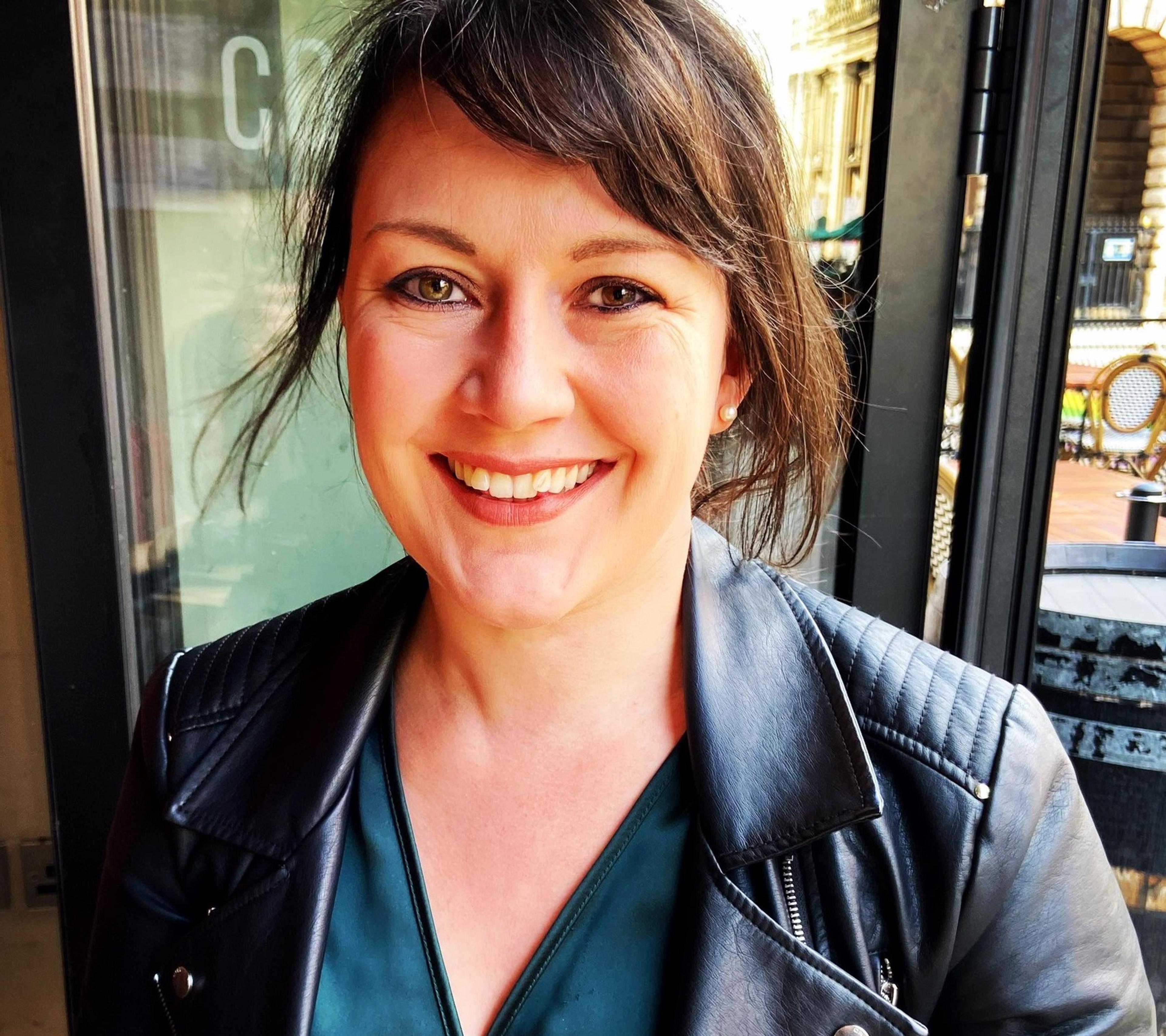 A smiling woman with dark brown hair wearing a black leather jacket and green top, standing in a well-lit outdoor café setting with glass doors and a city street in the background.
