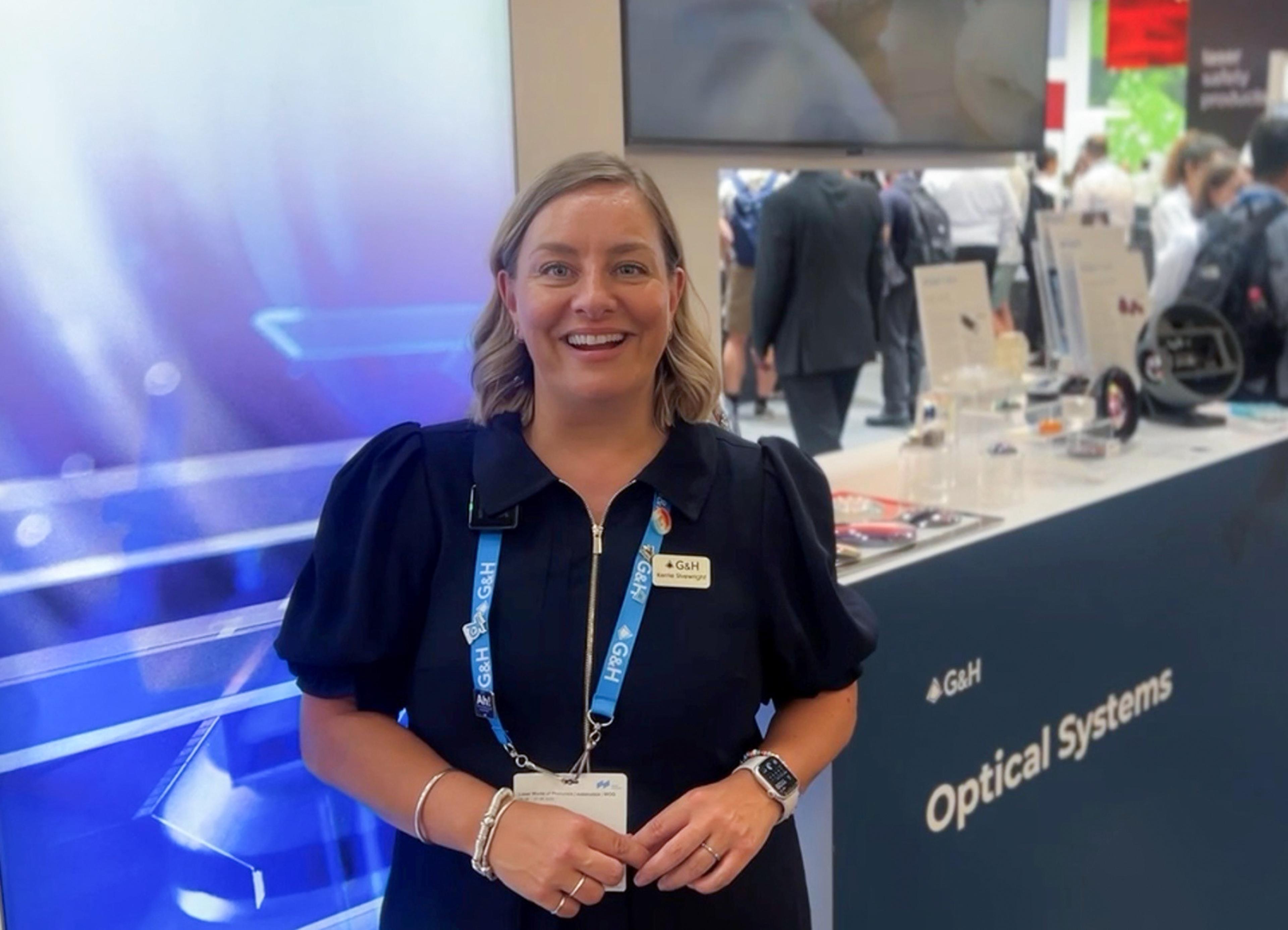Smiling G&H team member wearing a G&H lanyard and name badge stands at a trade show booth, with an “Optical Systems” display counter and product samples visible behind her.