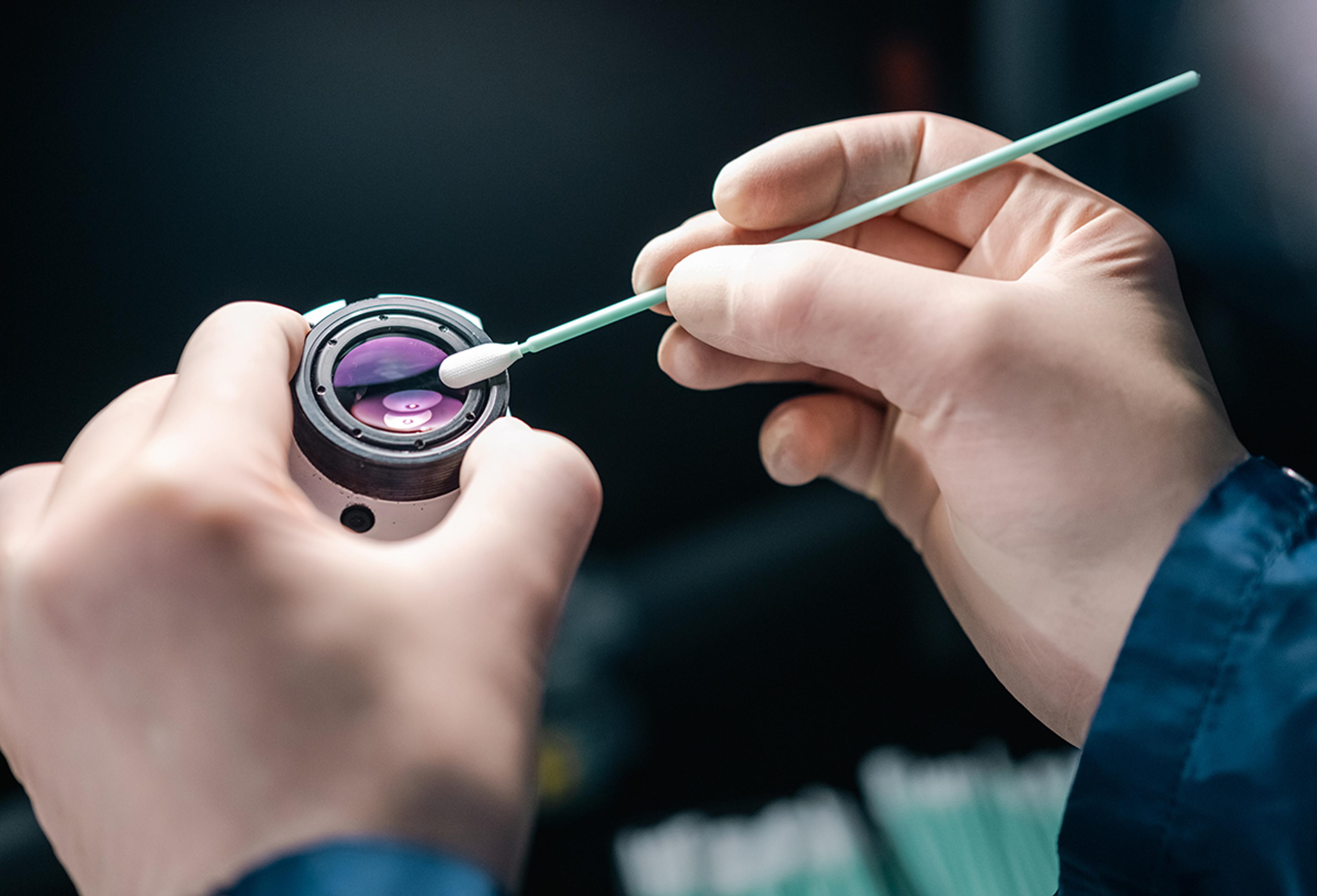 Hands cleaning a precision optical component with a swab in a controlled manufacturing setting.