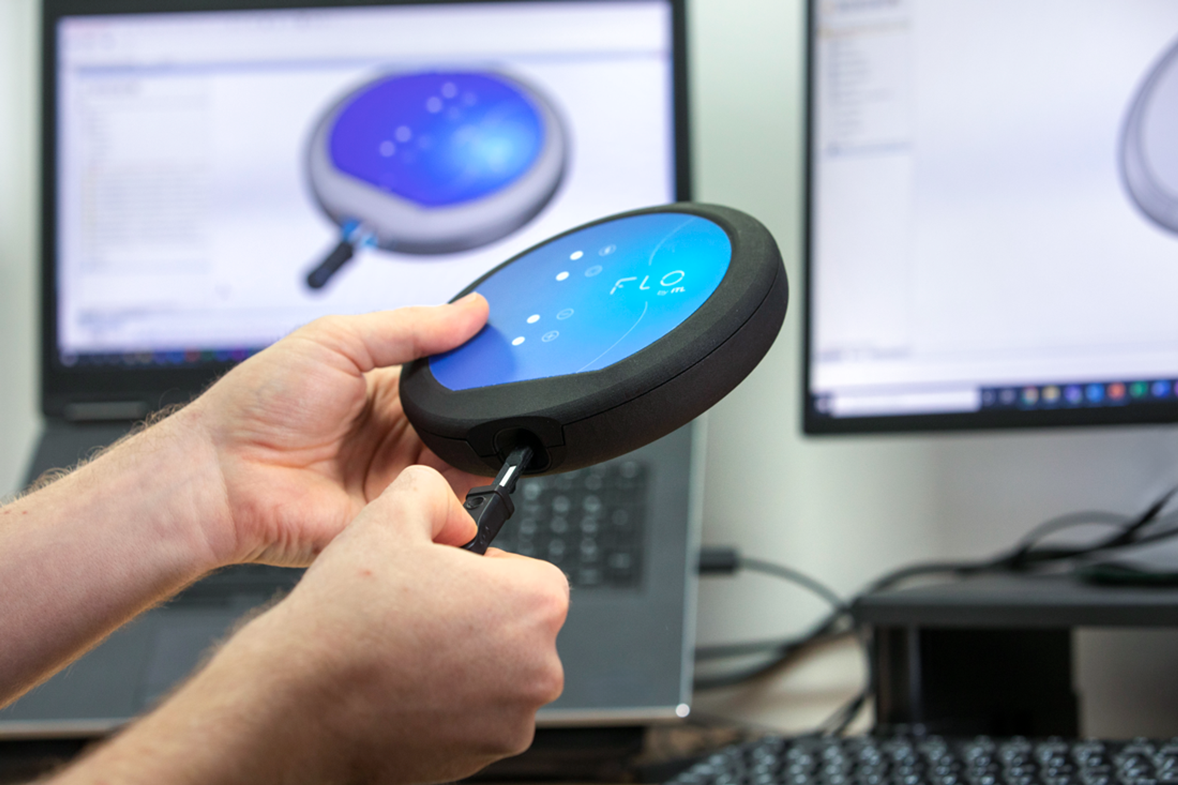 Close-up view of a person's hands connecting a cable to a round electronic device labeled "FLO by ITL." Behind the device, two computer screens display 3D CAD models of the same product, confirming the physical prototype's match with the digital design. The setup appears to be part of a product development or engineering workstation.