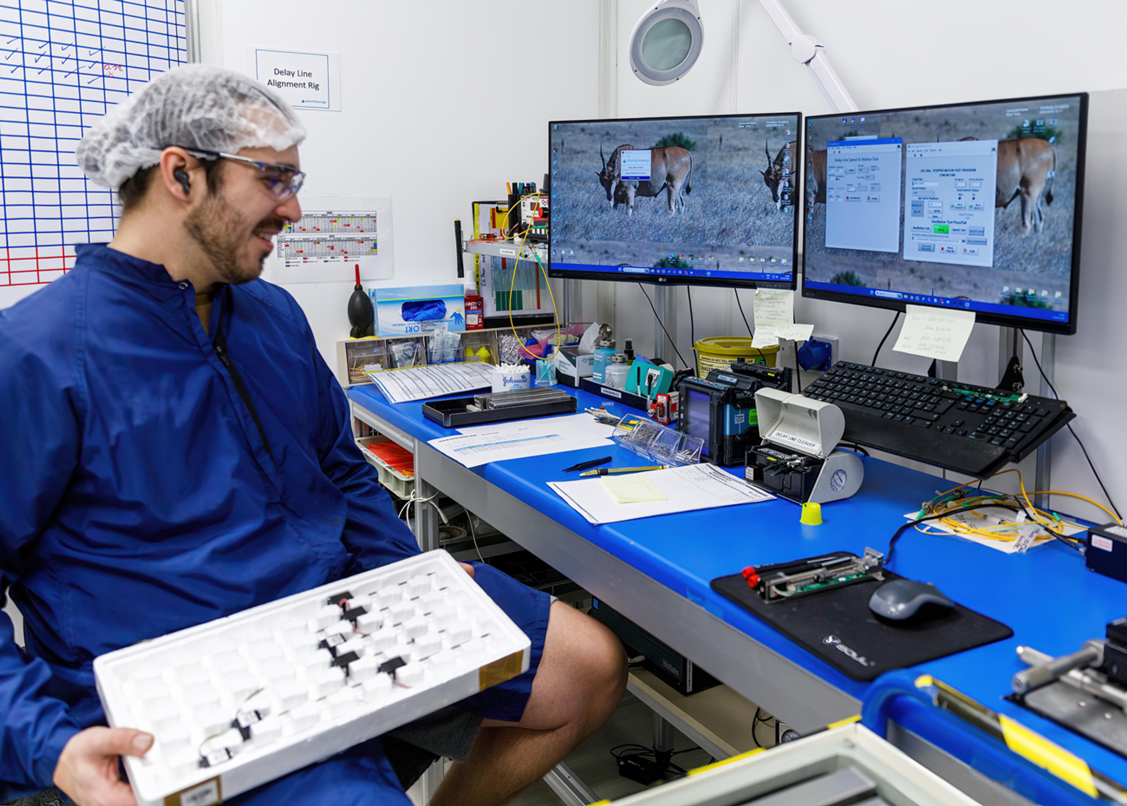 Technician in cleanroom clothing and a hairnet sits at a lab workstation with dual monitors, holding a tray of small optical components on a blue workbench.