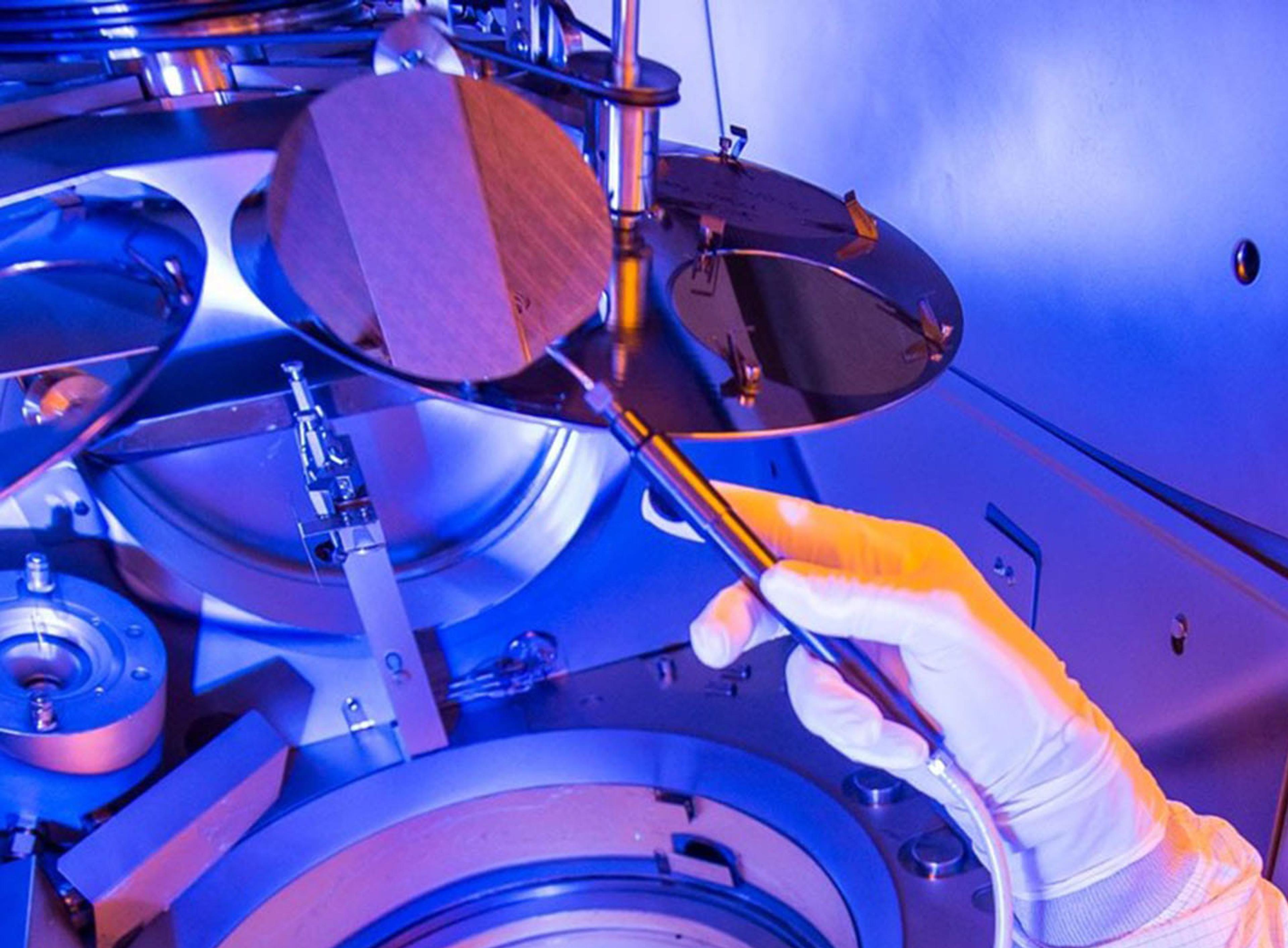 Gloved hand uses a vacuum wand to lift a thin circular wafer inside a blue-lit cleanroom processing chamber.