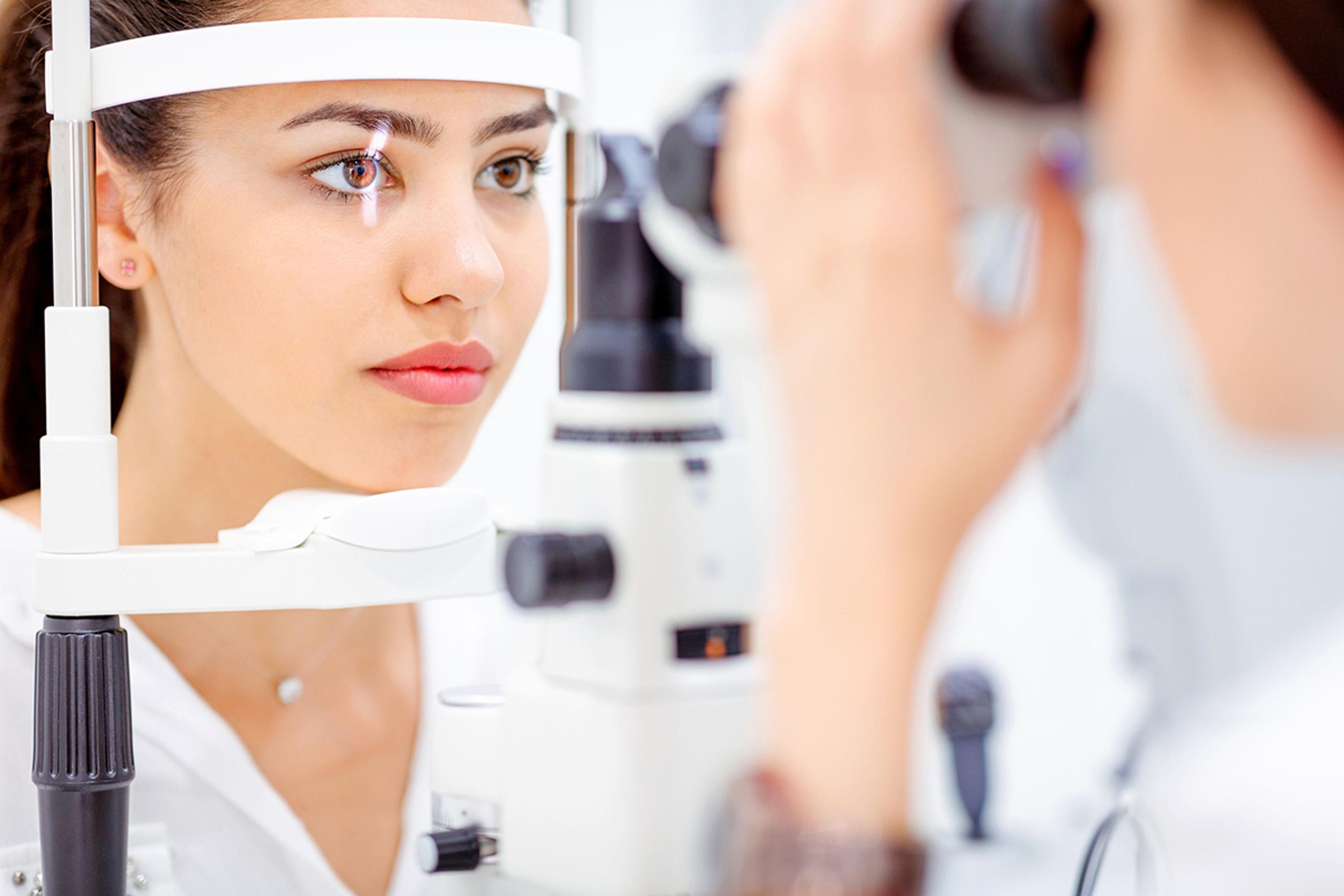 Optical coherent tomography (OCT) examination - A young woman undergoing an eye examination at an optometrist's clinic. She is seated with her chin resting on a support, looking into a slit lamp device. The examiner, slightly out of focus, is adjusting the equipment to inspect her eye. The setting is clean and brightly lit, emphasizing the clinical environment.
