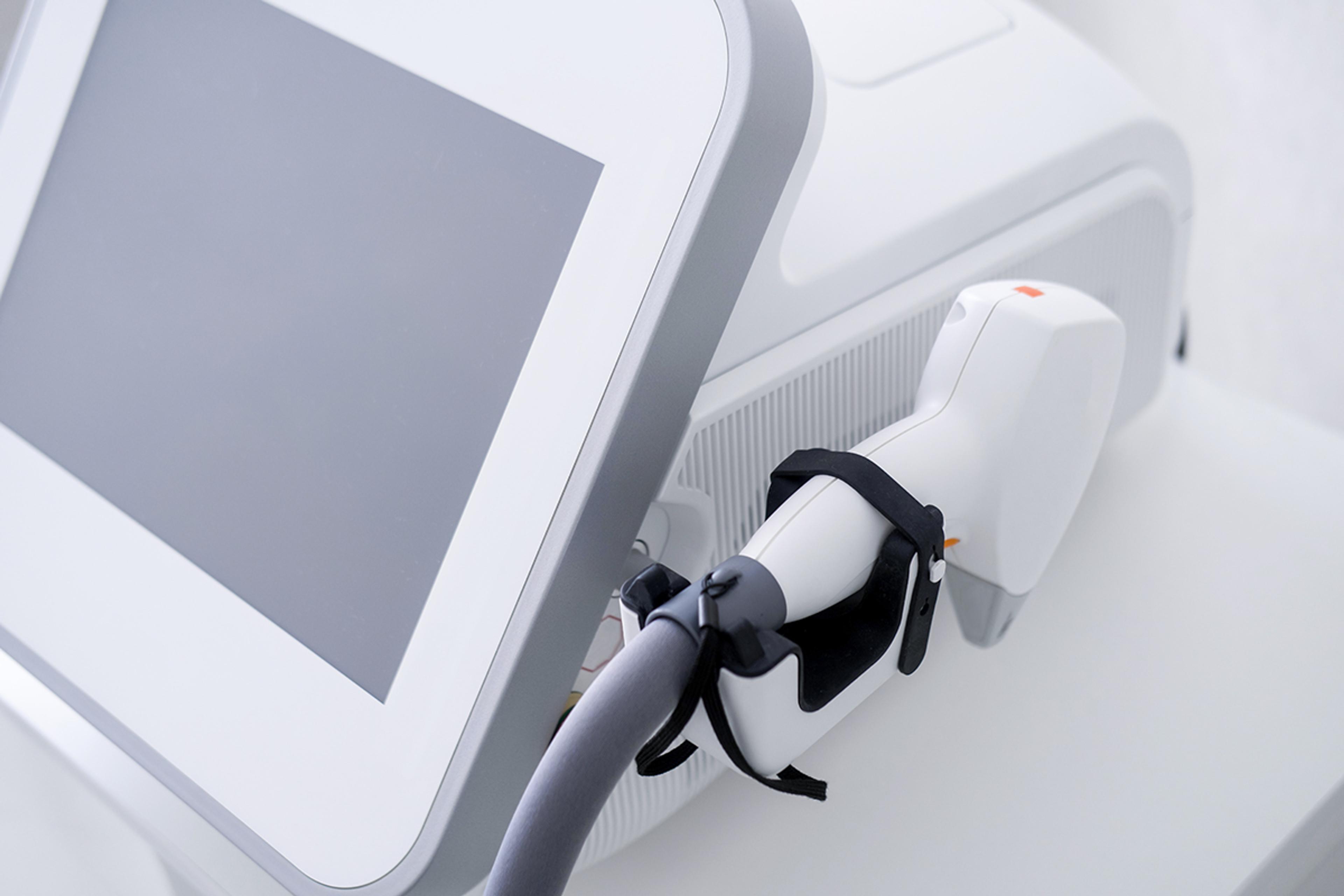 Handheld aesthetic/medical laser treatment applicator docked in its cradle beside a touchscreen console on a white clinical device.