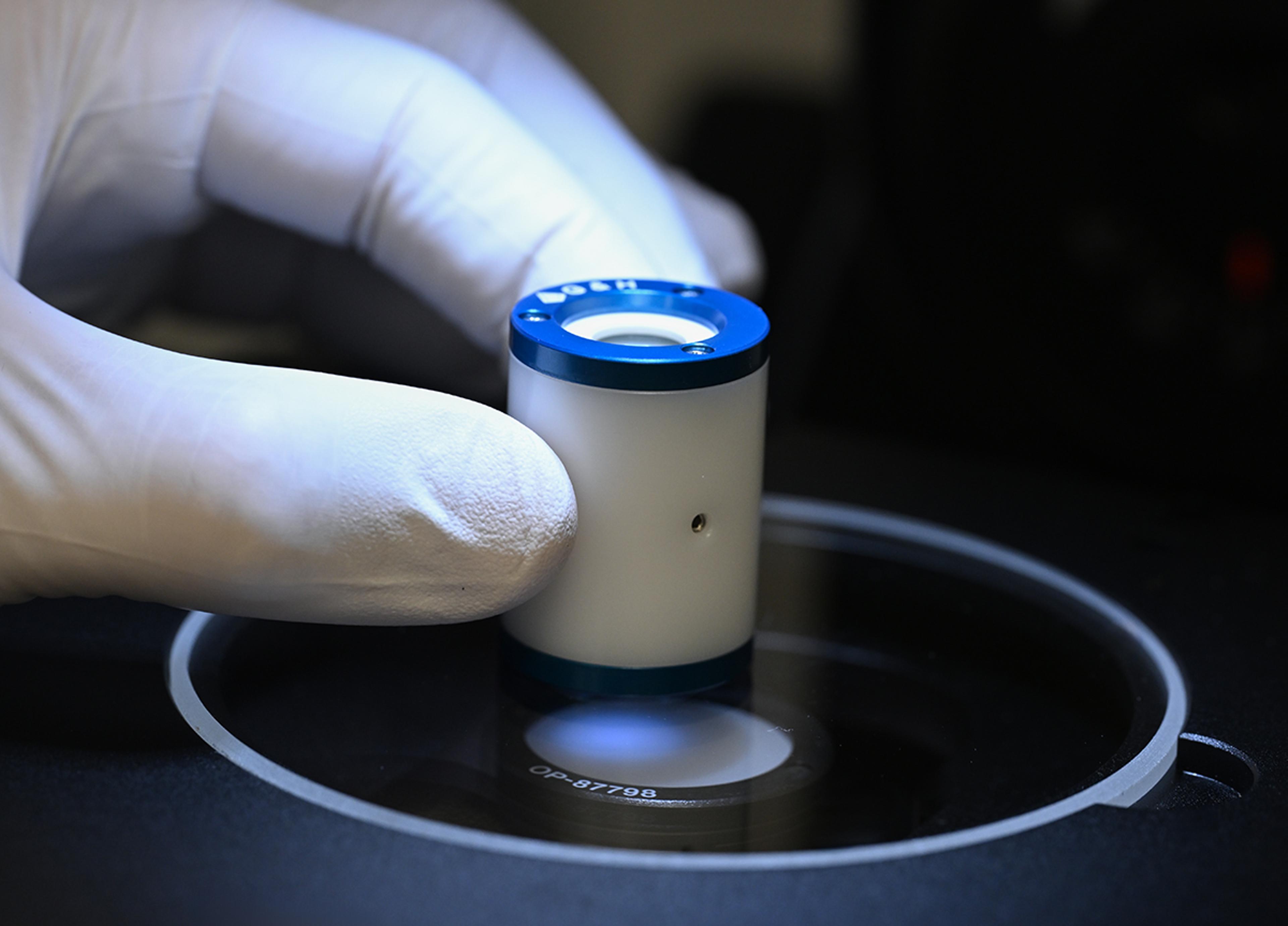 Close-up of a gloved hand positioning a Pockels cell cylindrical optical component with a blue top and white body, marked with the G&H logo, onto a precision inspection platform.