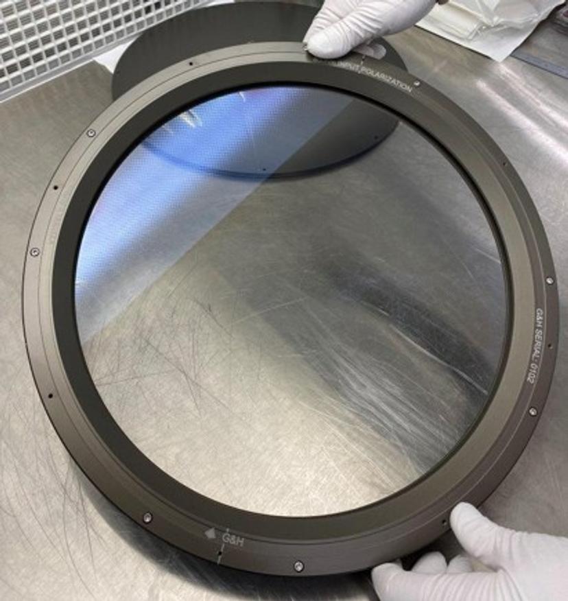 Gloved technician holding a large precision optical waveplate in a metal mounting ring, manufactured by G&H, on a cleanroom work surface.