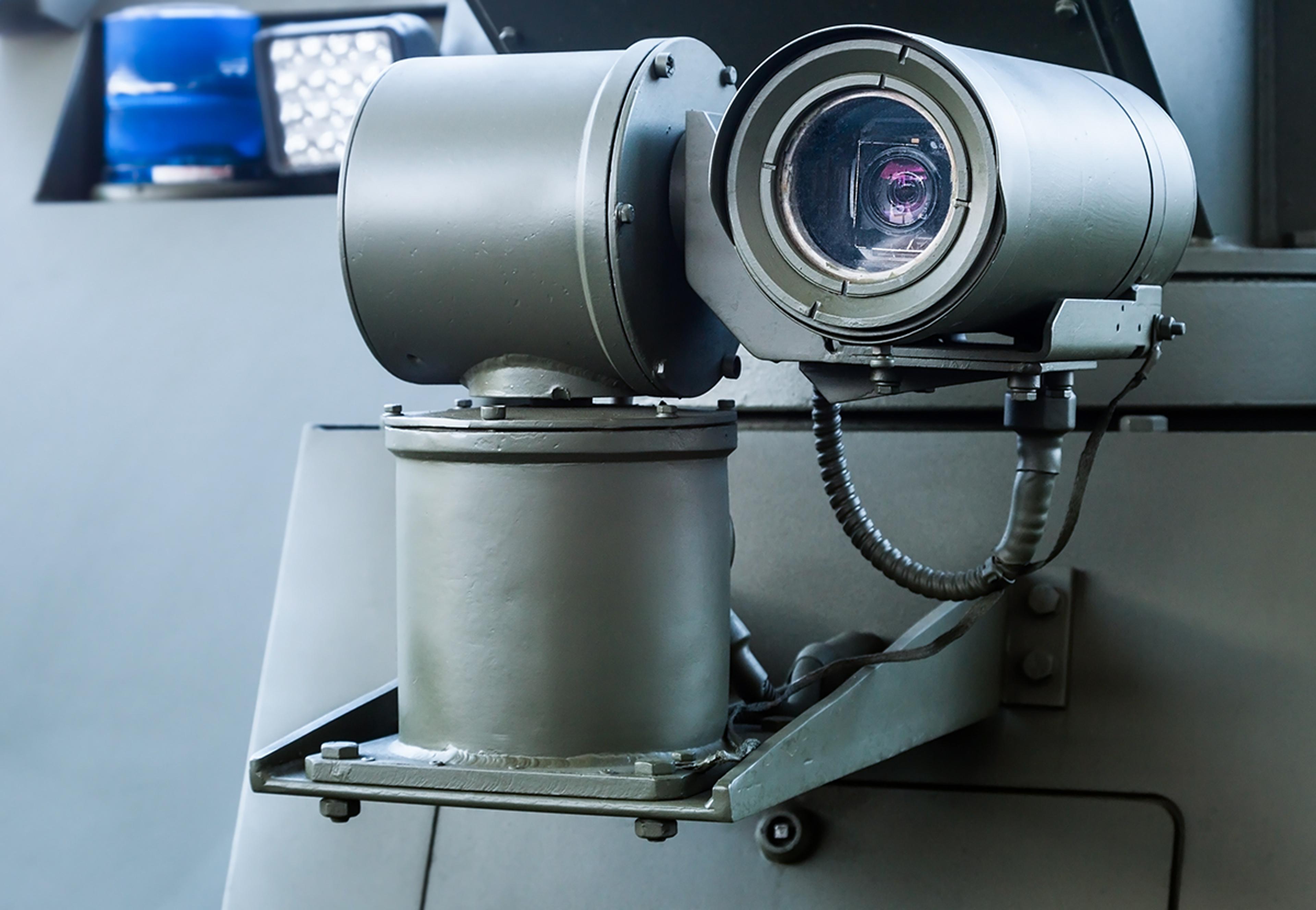 Close-up of an armoured vehicle’s turret-mounted electro-optical camera/sight unit, with a large circular lens in a protective housing and cabling attached.
