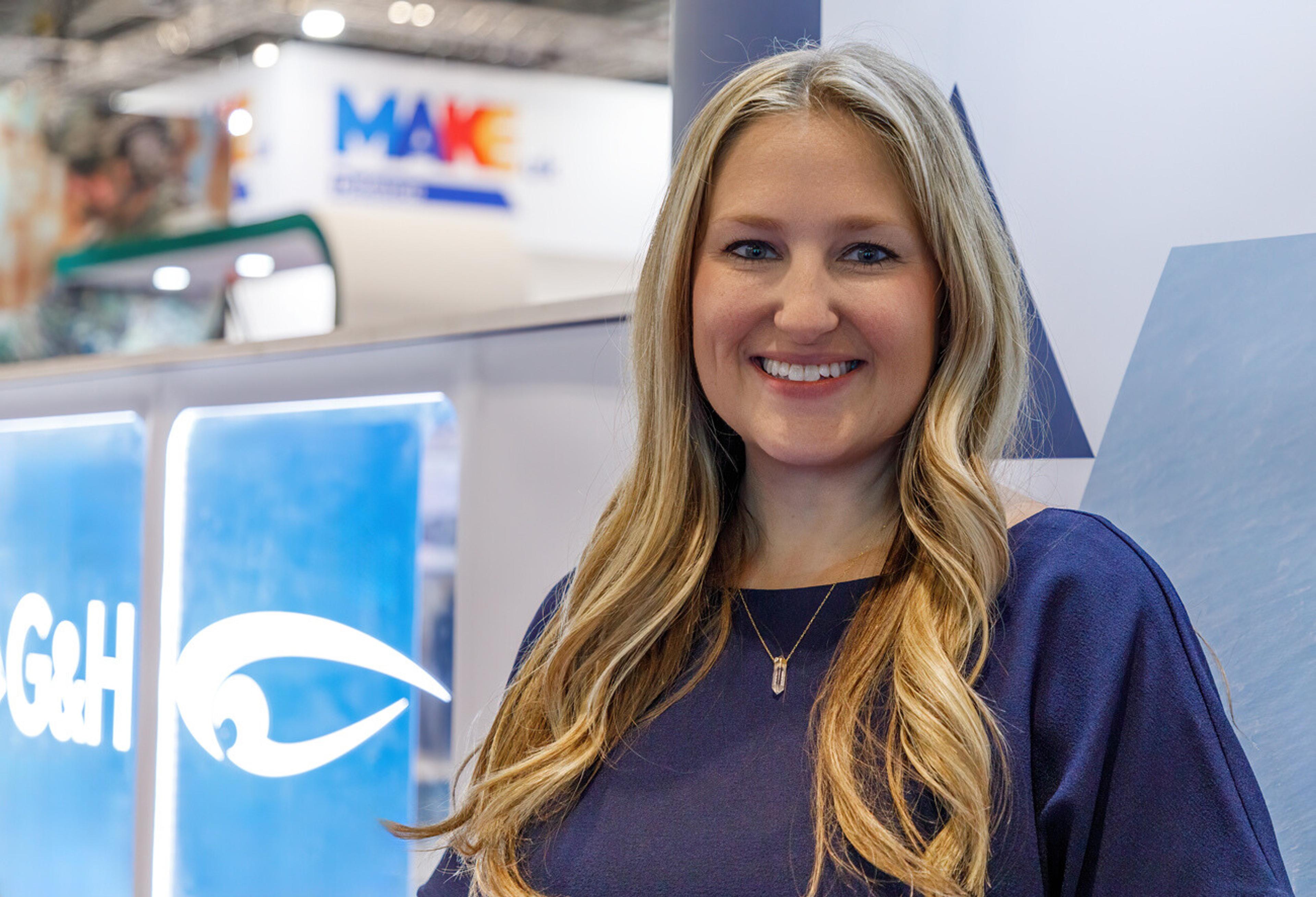 Smiling woman standing at a trade show booth with G&H branding and signage visible in the background.