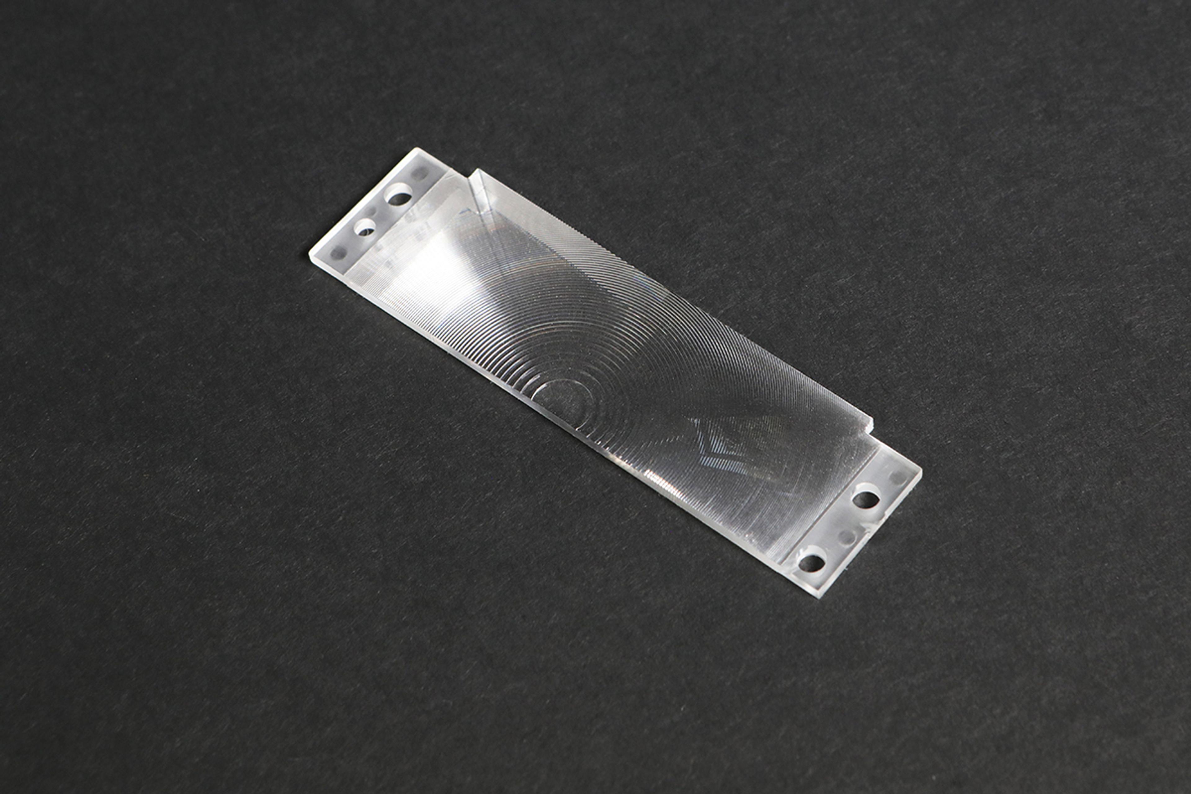 Close-up of a precision-engineered transparent optical component with concentric circular grooves, possibly a Fresnel lens or diffractive optical element, placed on a dark surface. The component has mounting holes at both ends.