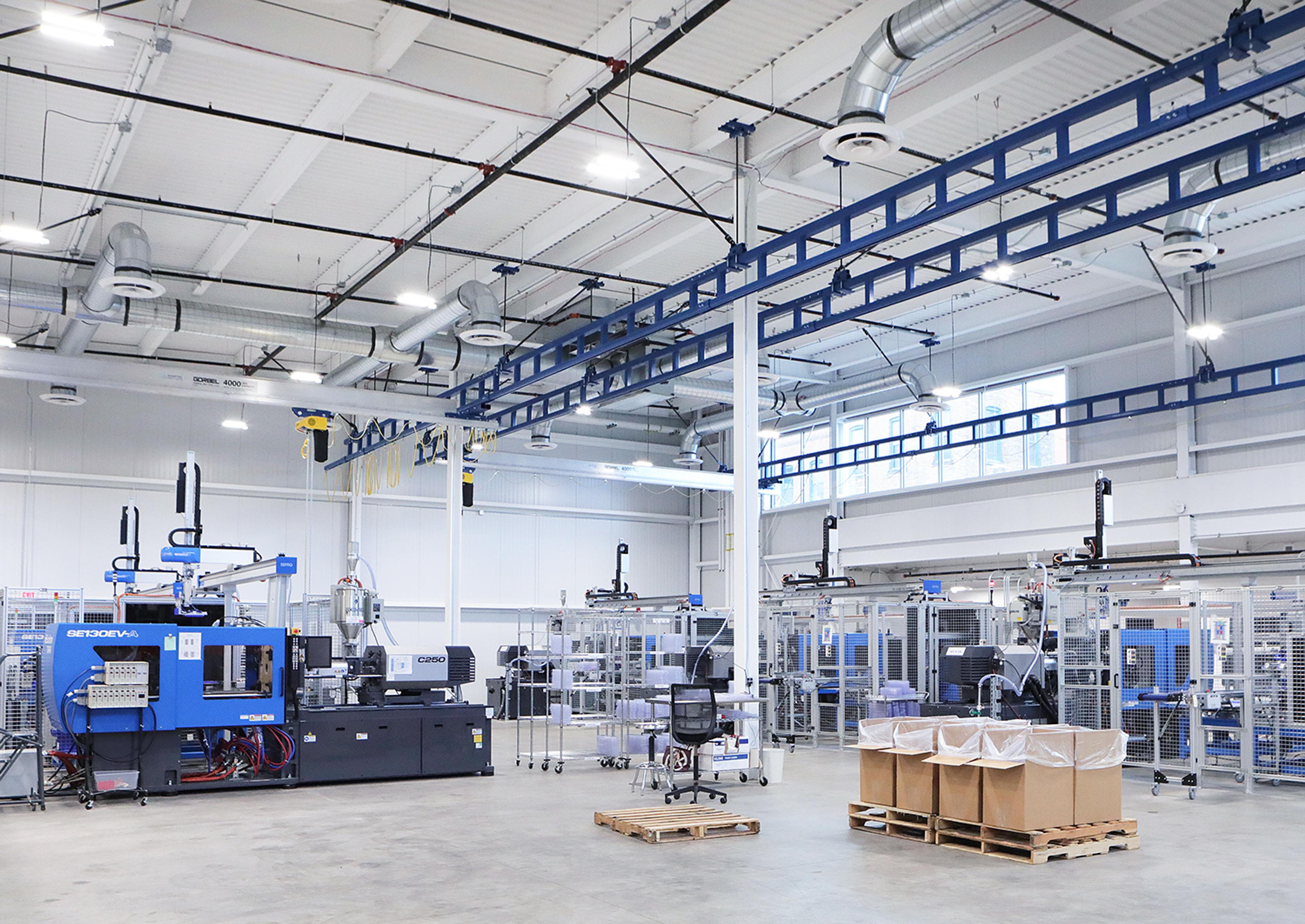 Inside our Rochester site, spacious and well-lit modern manufacturing facility featuring a series of injection molding machines and robotic automation systems. The high-ceilinged room includes visible ductwork, bright overhead lighting, and organized workstations. Large cardboard bins on pallets and a few office chairs are positioned on the clean concrete floor, contributing to a streamlined and efficient industrial environment
