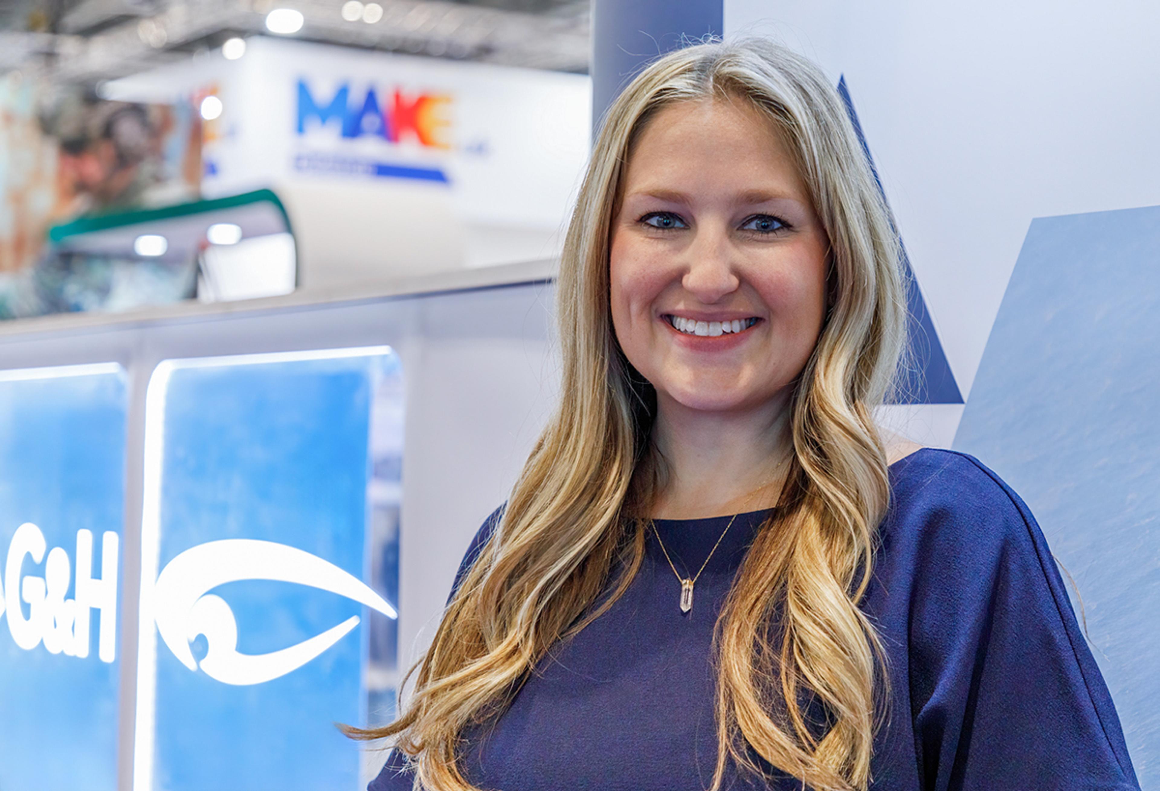 Smiling woman standing at a trade show booth with G&H branding and signage visible in the background.