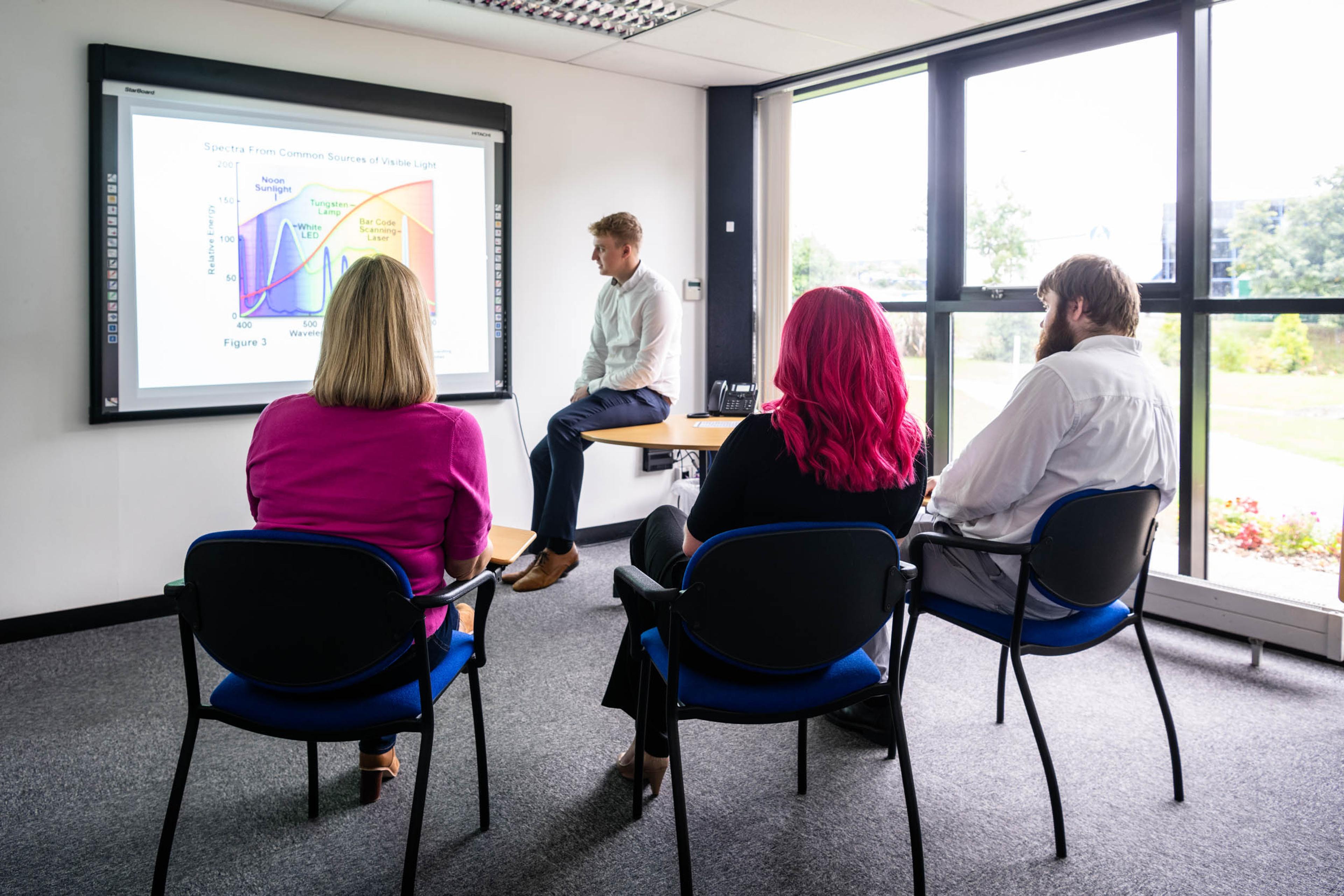 A group of four people are in a meeting room attending a presentation. Three participants are seated facing a screen, while the presenter, dressed in a white shirt, sits on the edge of a desk near a projection showing a visible light spectrum chart. Large windows let in natural light, creating a bright and modern setting.