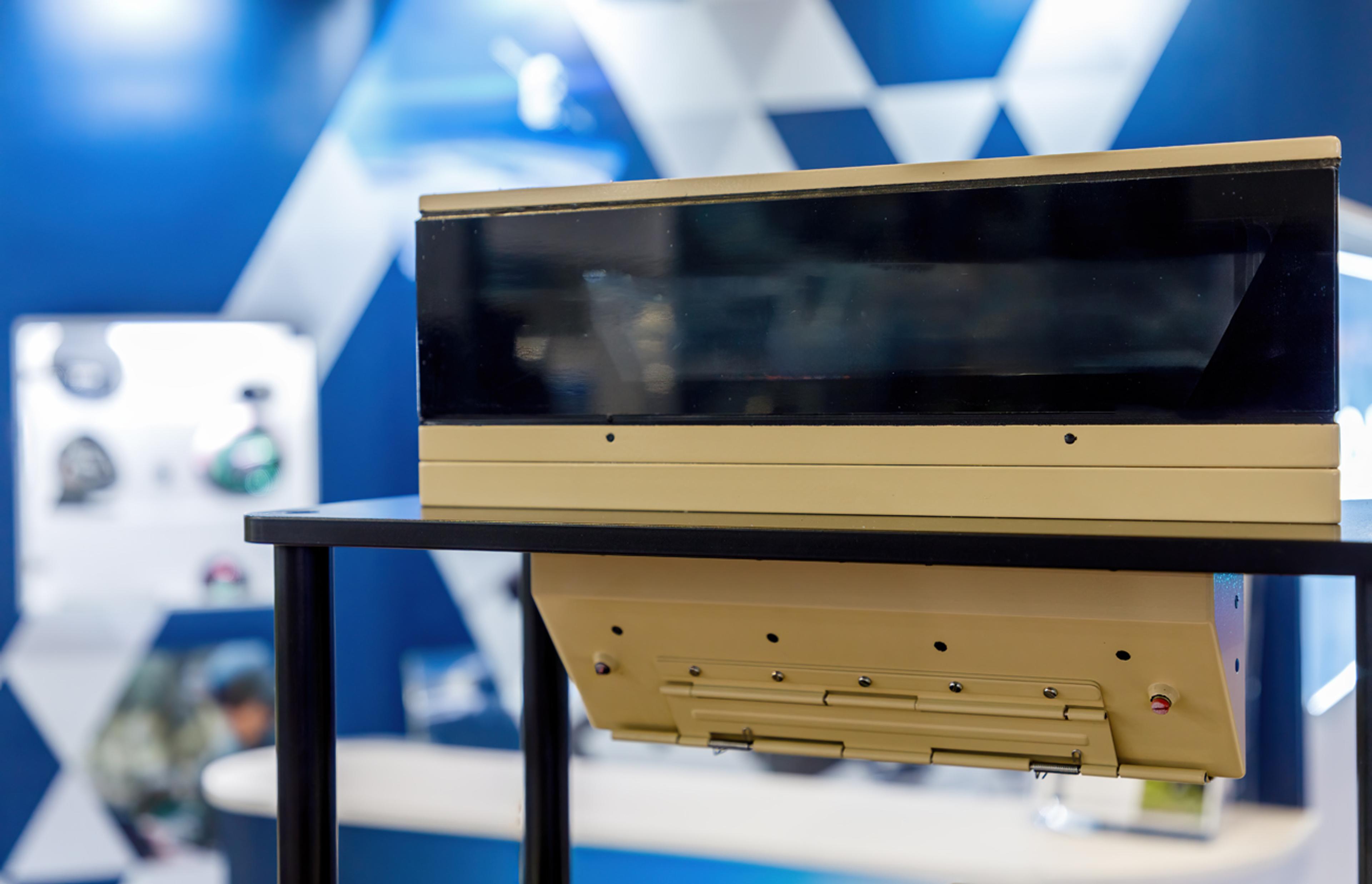 Armoured vehicle periscope or vision module displayed on a stand, featuring a wide protective viewing window and ruggedised housing in a tan military finish.