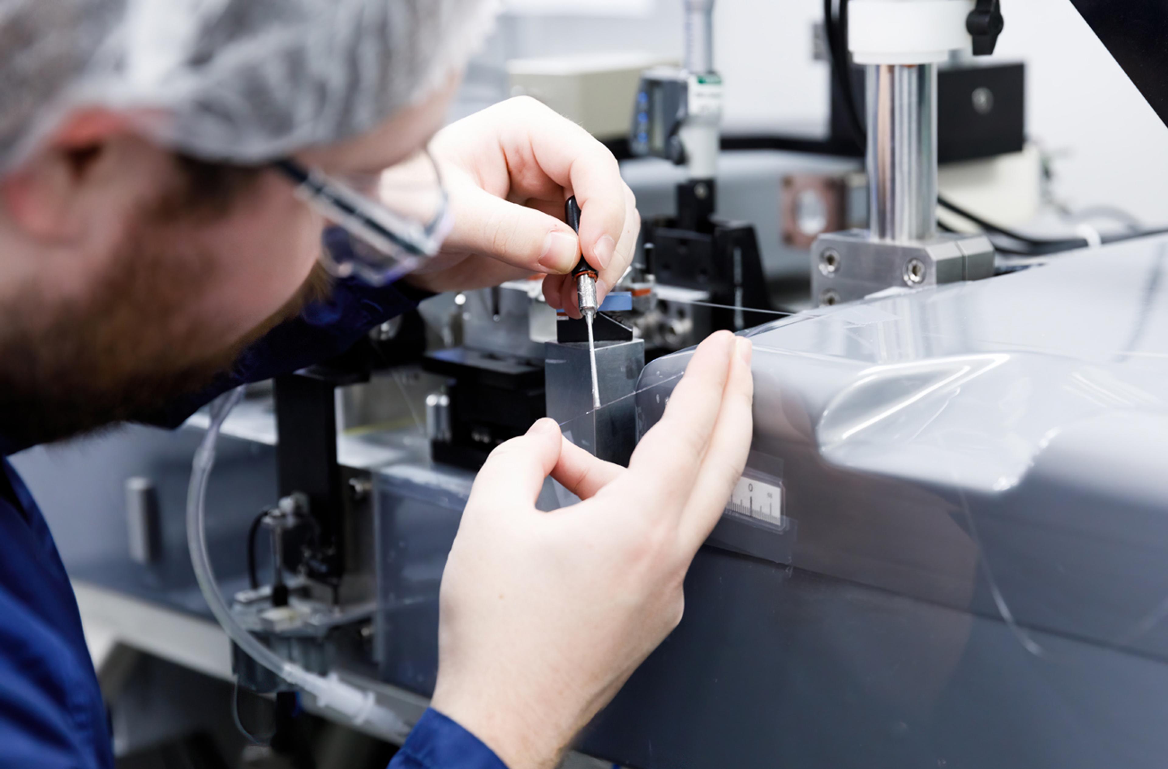Close-up of a technician wearing safety glasses using a hand tool to work on a fine fibre optic within a precision assembly fixture, behind a clear protective shield.