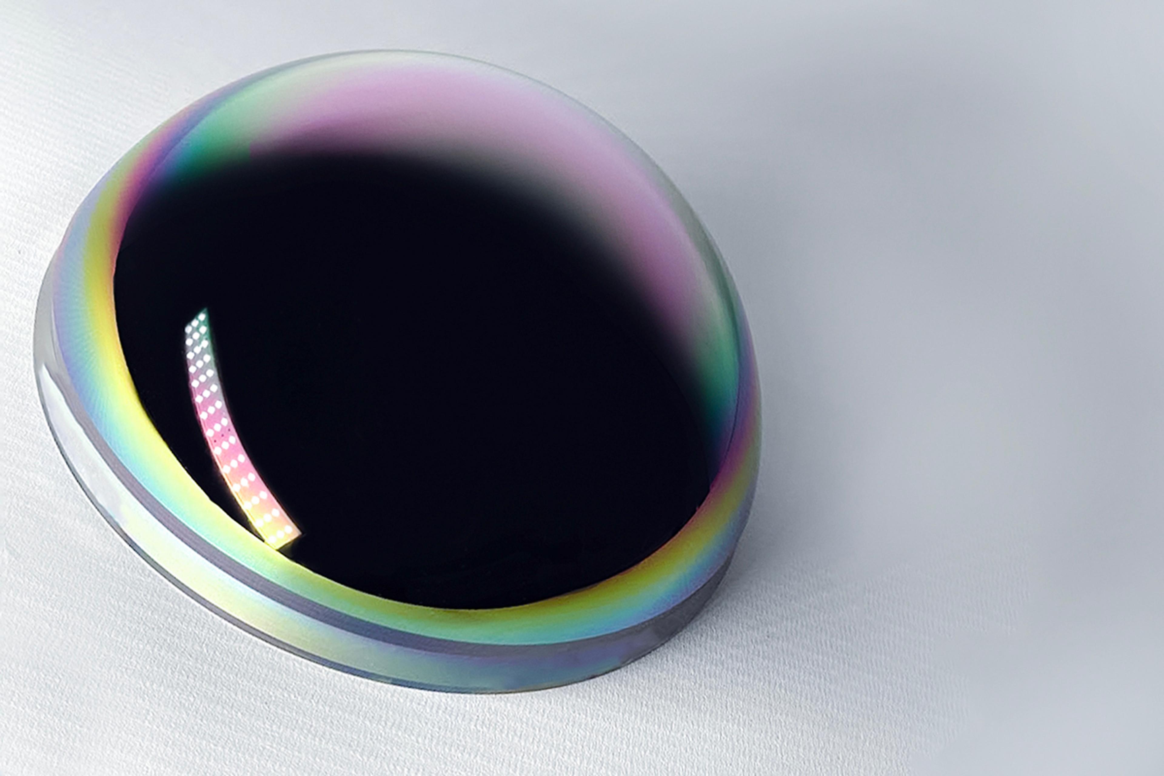 A single high-curvature Germanium lens with a reflective black surface and vivid rainbow-colored anti-reflective coating around the edge, positioned on a soft gray background. The coating indicates optimization for infrared transmission, commonly used in MWIR or LWIR optical systems.
