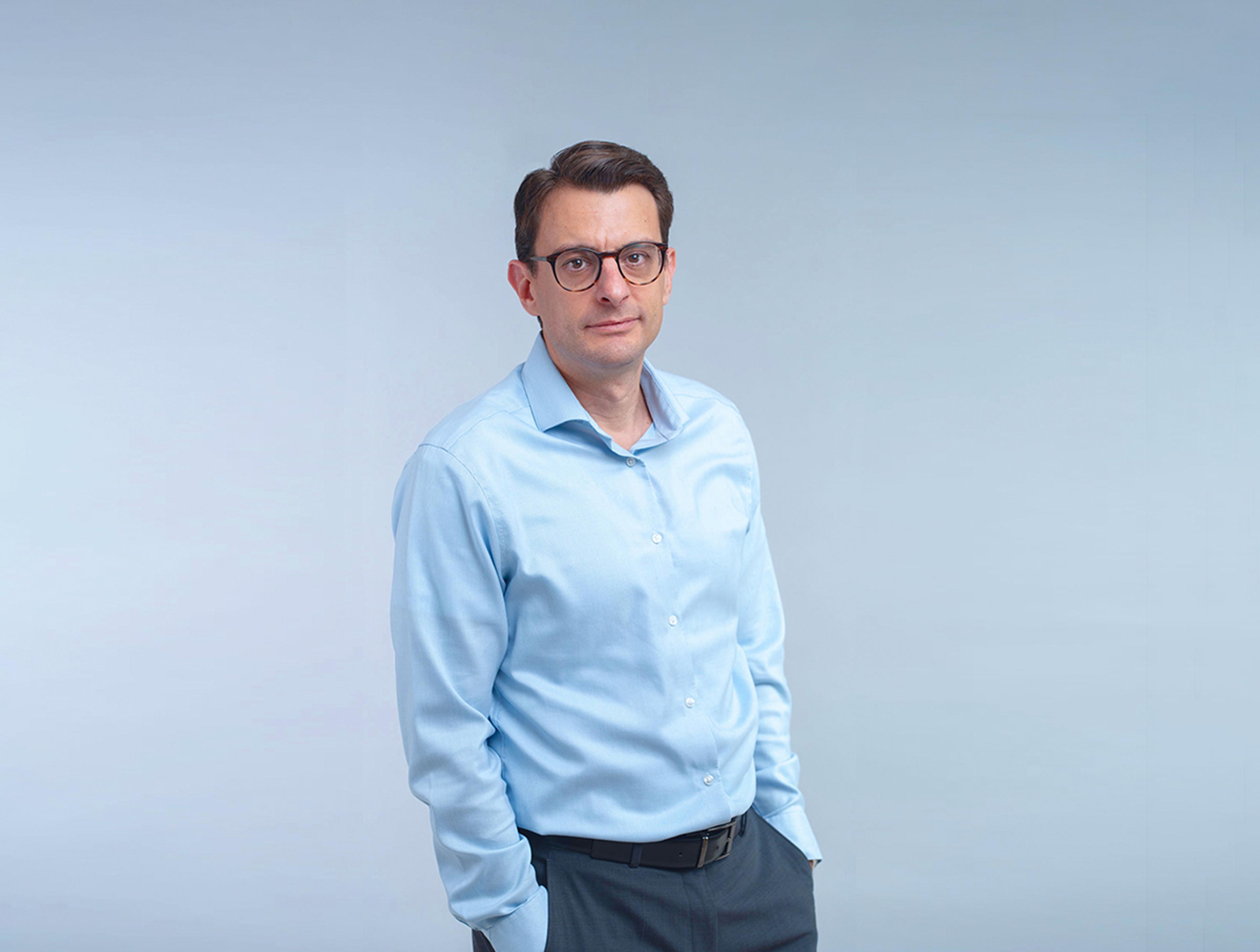Portrait of a man wearing glasses and a light blue shirt, standing with his hands in his pockets against a pale blue background.