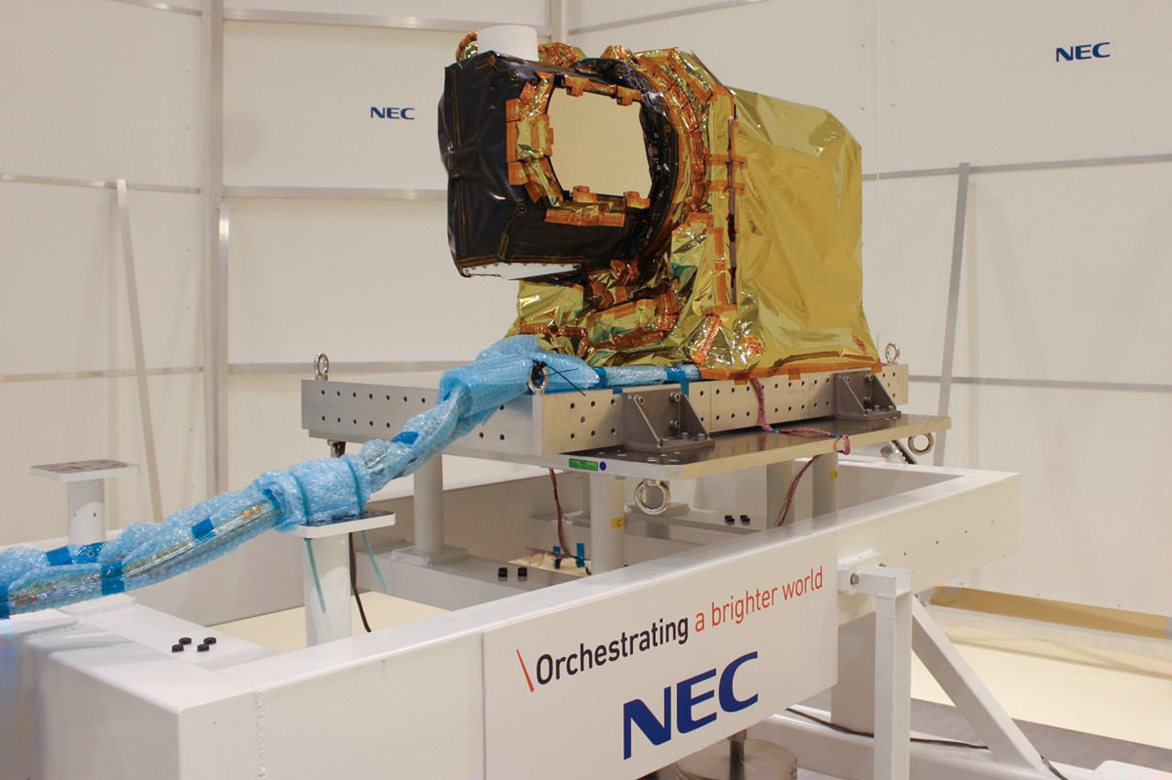 Laser communication terminal for geostationary satellites, A gold-foil-wrapped satellite optical payload is mounted on a testing platform in a cleanroom. The device, part of NEC's space technology, features a black aperture and various cables wrapped in blue protective material. The platform is labeled with NEC's slogan "Orchestrating a brighter world." The white walls of the cleanroom bear the NEC logo in multiple places