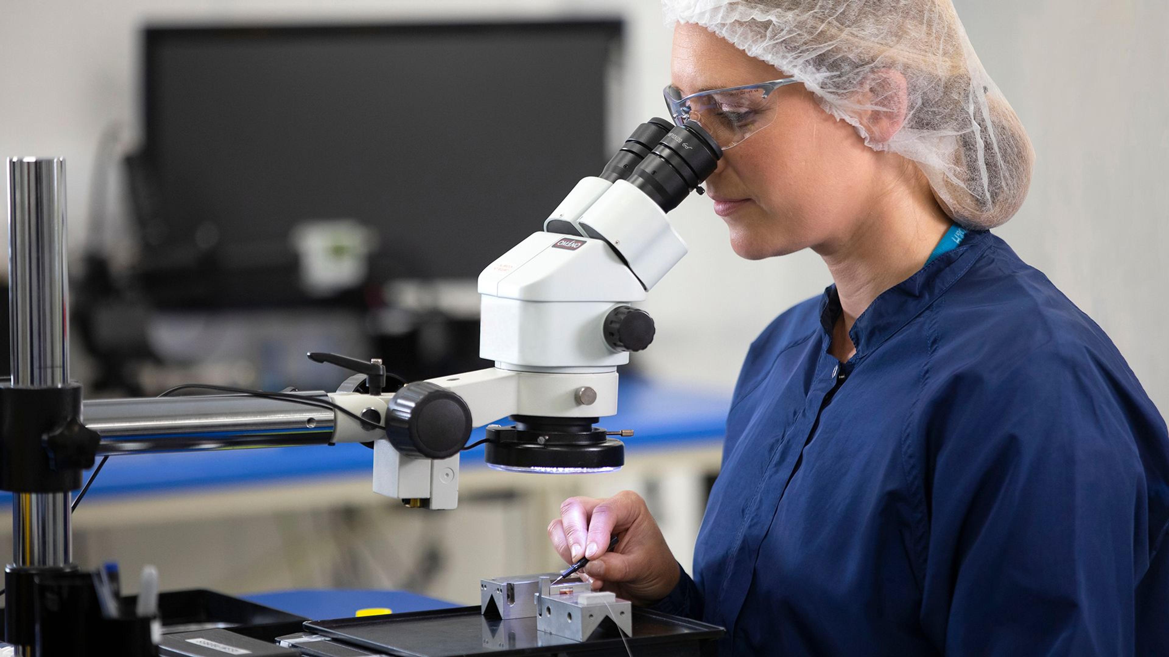 A technician in a cleanroom gown, hair net, and safety glasses uses a microscope to inspect a precision optical component on a metal fixture in a laboratory setting.