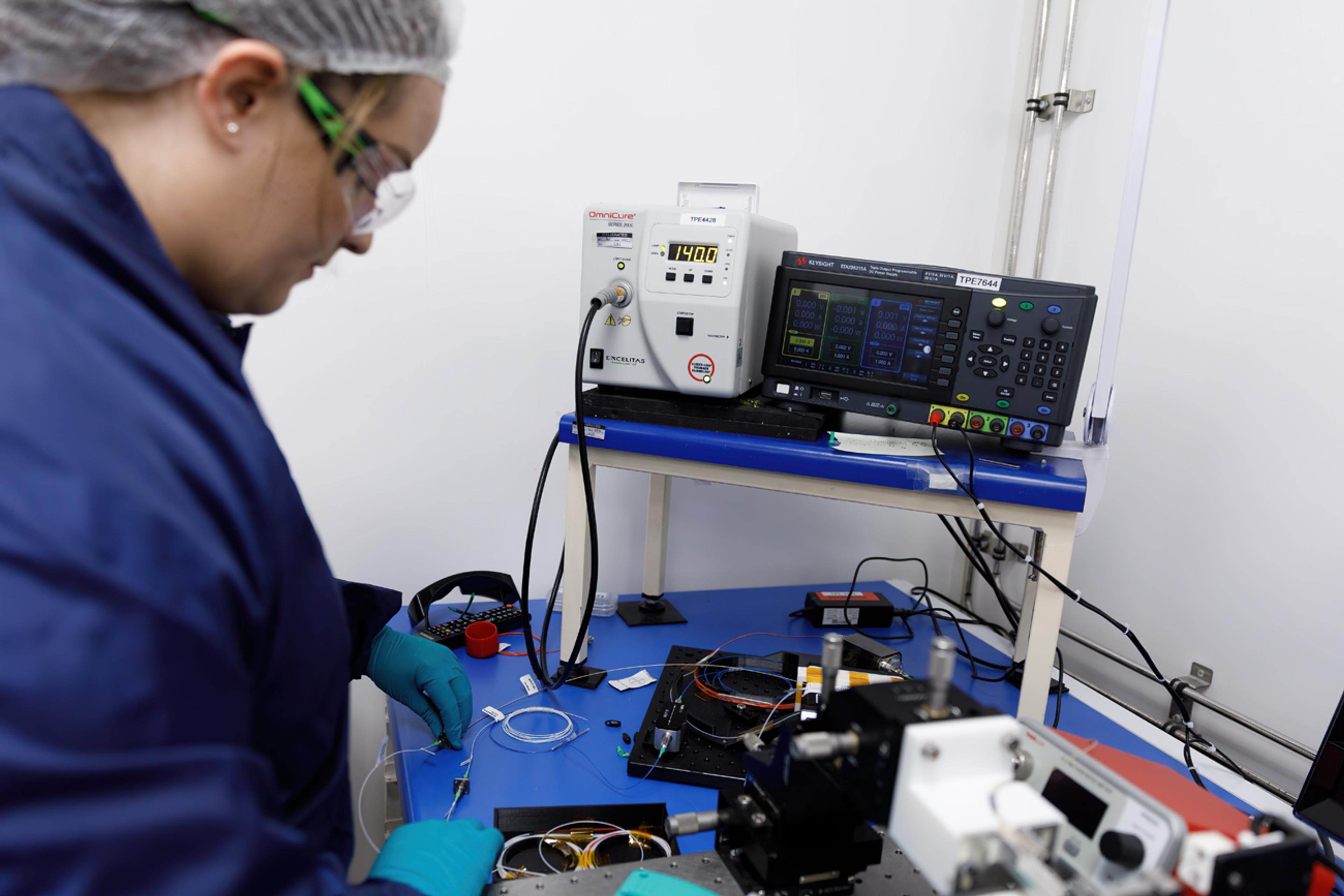 Technician wearing safety glasses and blue gloves works at an optical test bench, adjusting fibre-optic components beside lab instruments and a laser/measurement control unit.