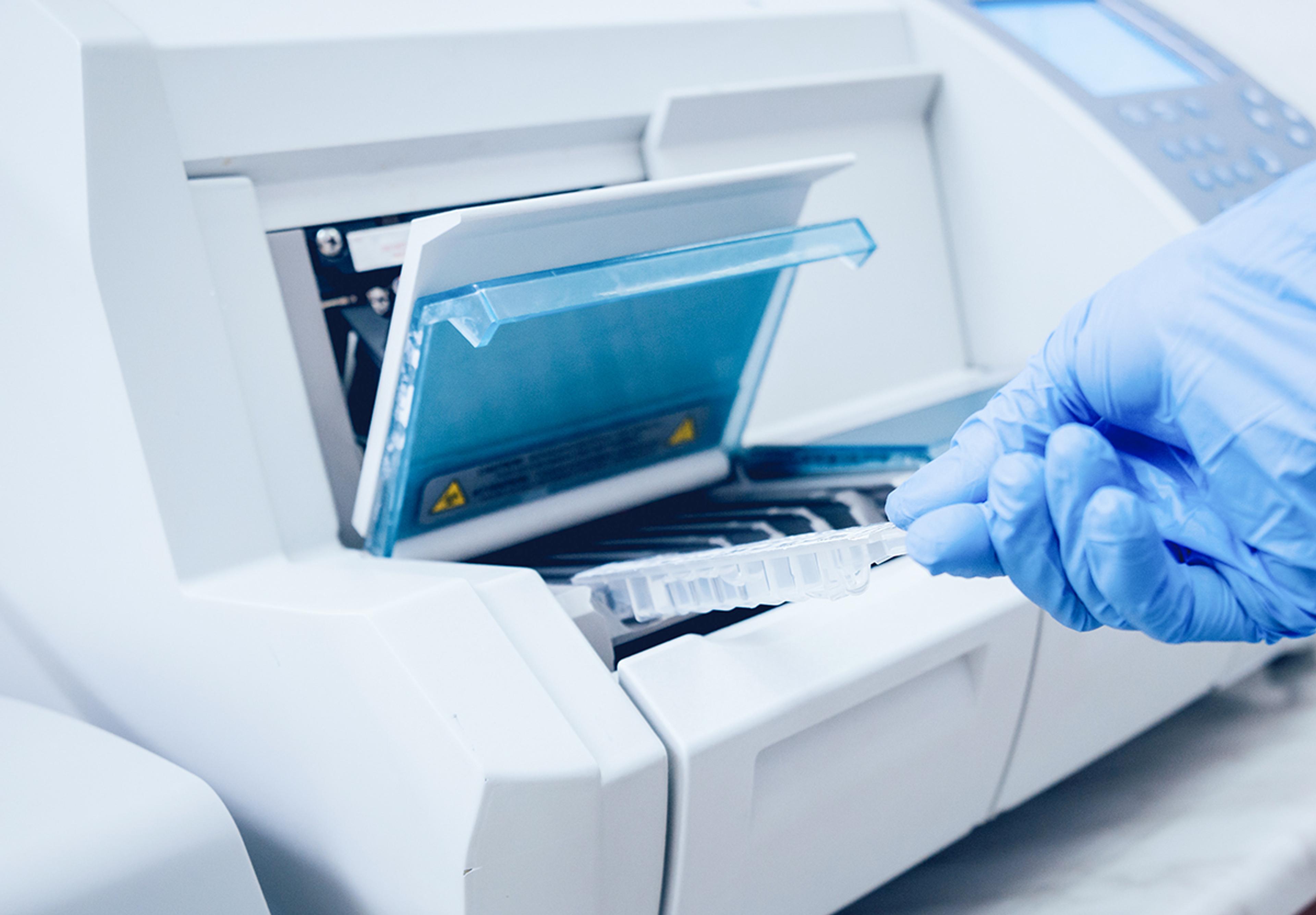 Gloved hand loading a sample tray into a laboratory analyser with the instrument lid open.