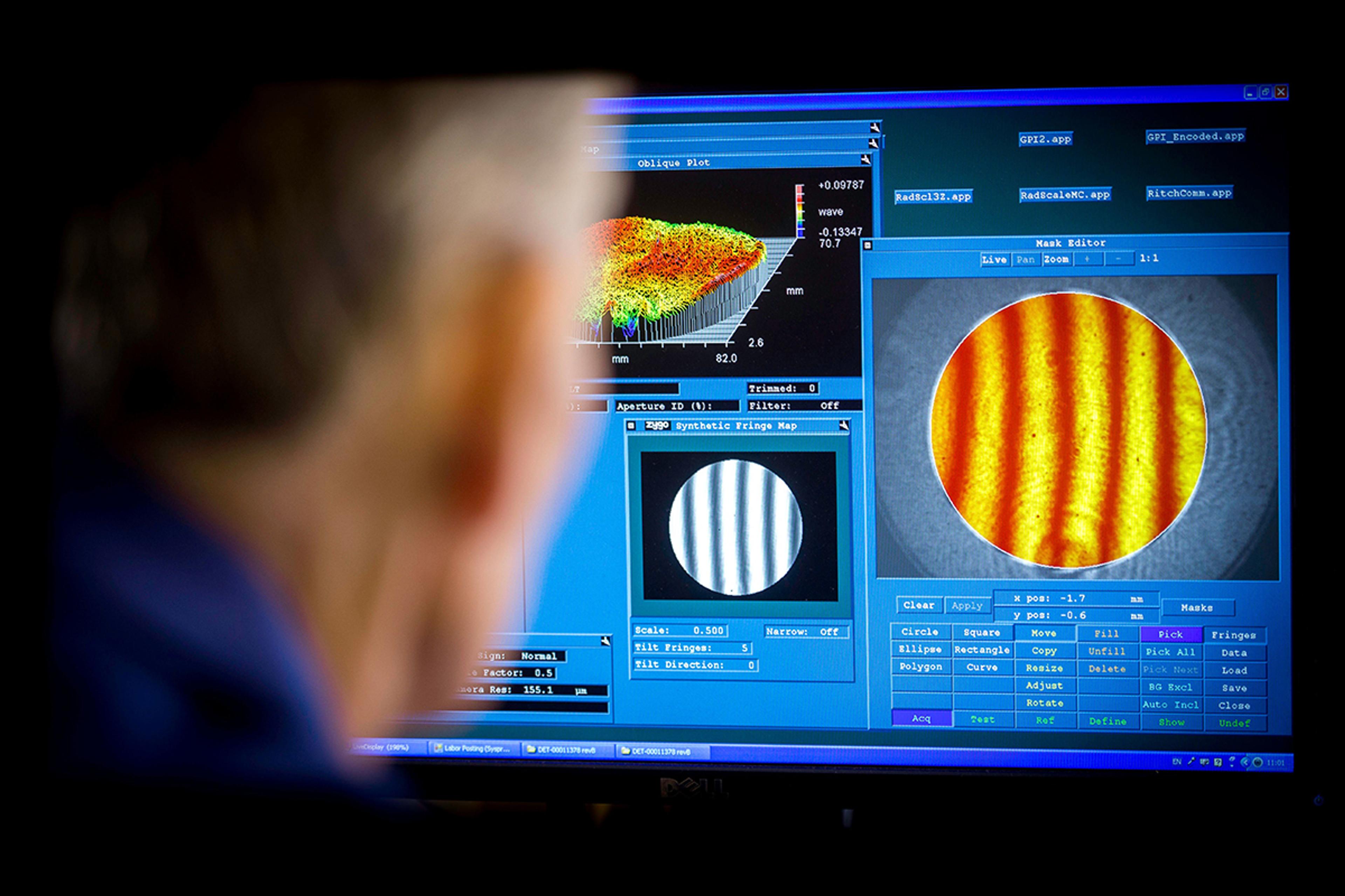 Technician controlling the profile of a precision optics component at G&H Ilminster - A person closely observes a computer screen displaying optical metrology data. The monitor shows various analysis windows including a 3D surface plot, synthetic fringe patterns, and a detailed circular interferogram with orange and yellow fringes. The scene suggests precision optical inspection or surface quality evaluation.