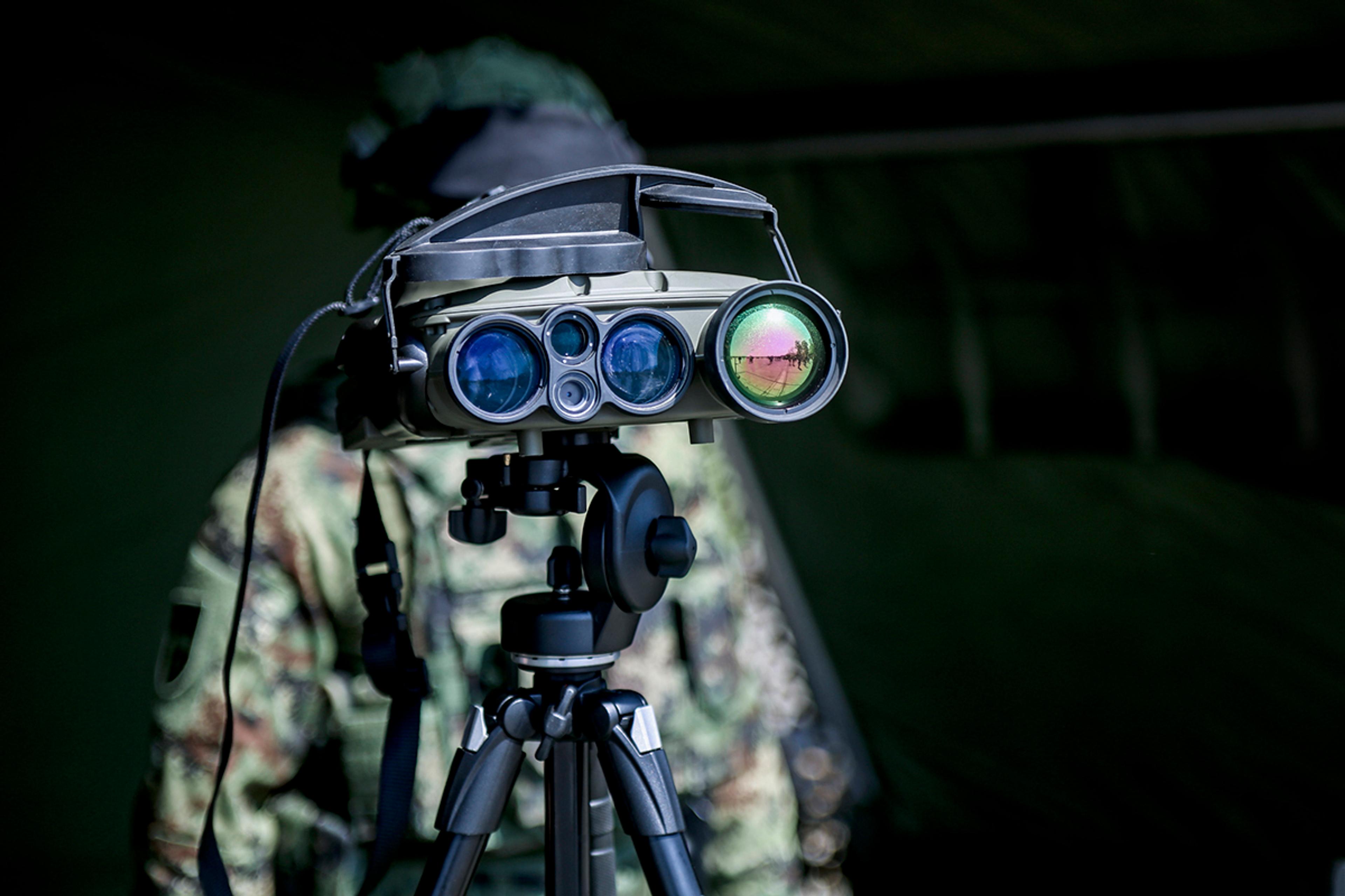 Tripod-mounted multi-sensor optical surveillance system with multiple precision lenses, operated by a person in camouflage.
