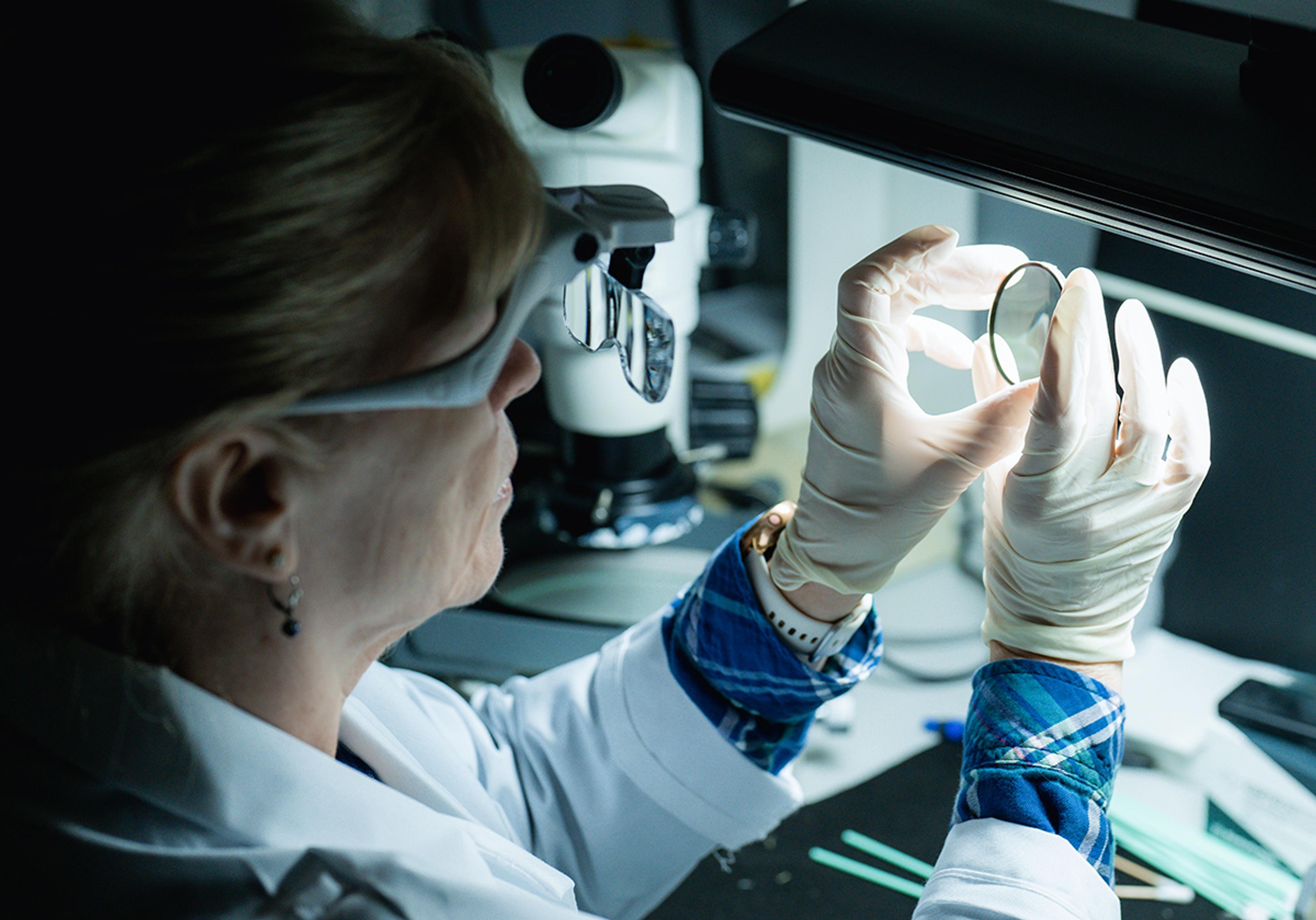 Female technician wearing magnifying eyewear and gloves inspects an optical lens under a bright work light, with a microscope and precision tools in the background